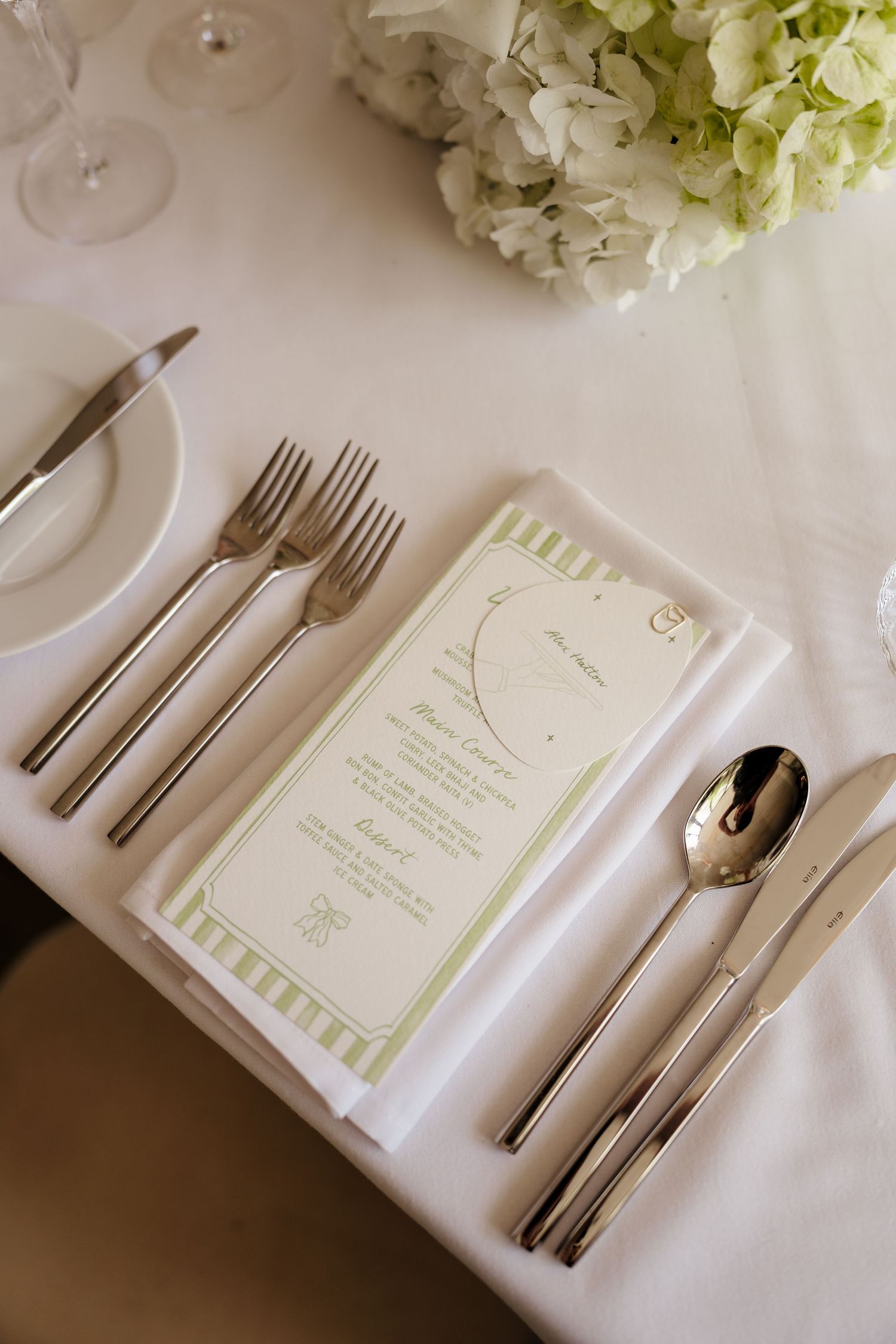Wedding reception place setting with green striped menu card, silverware, and white hydrangea centerpiece.