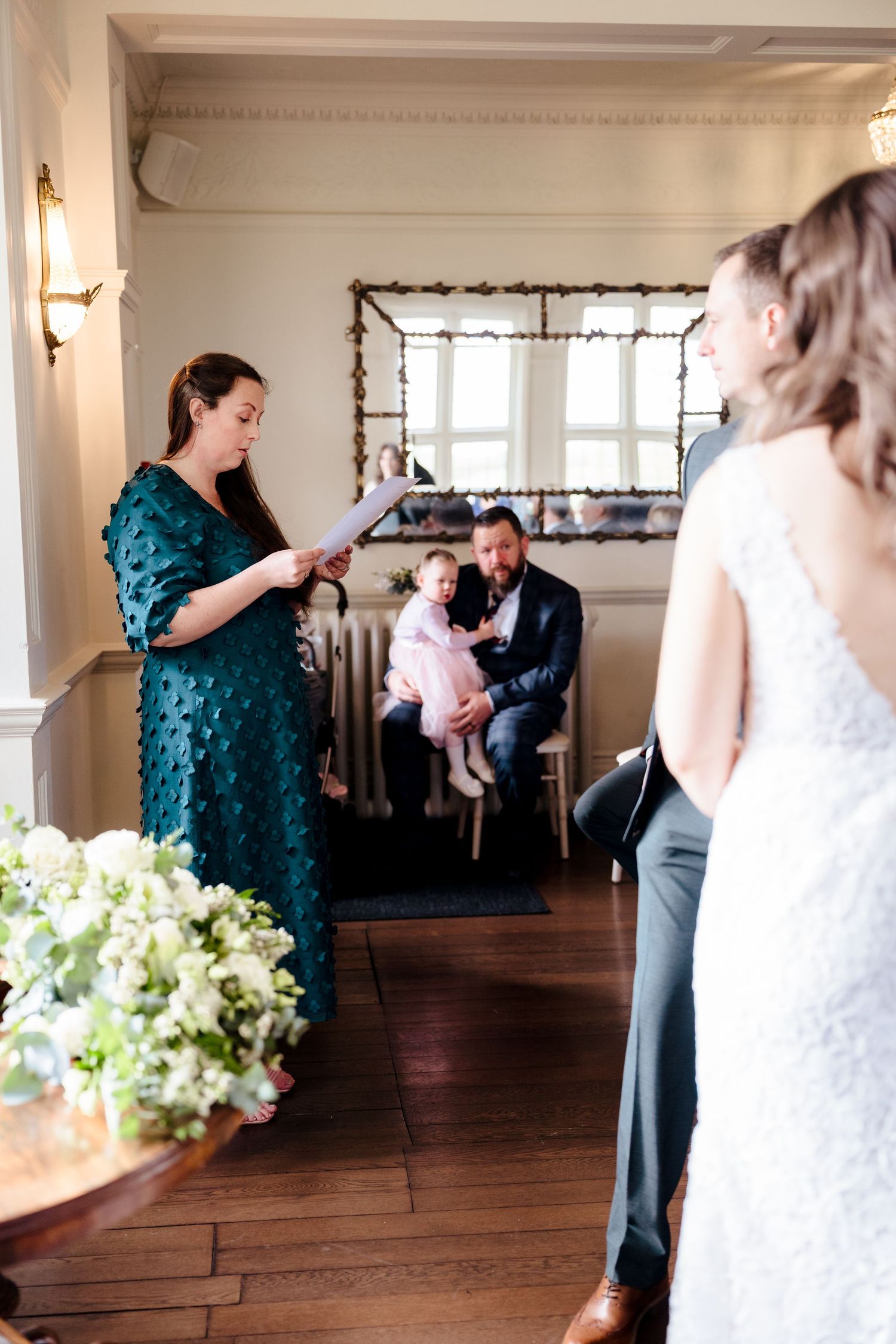Wedding ceremony reading by a guest in teal dress as groom holds child in the background.