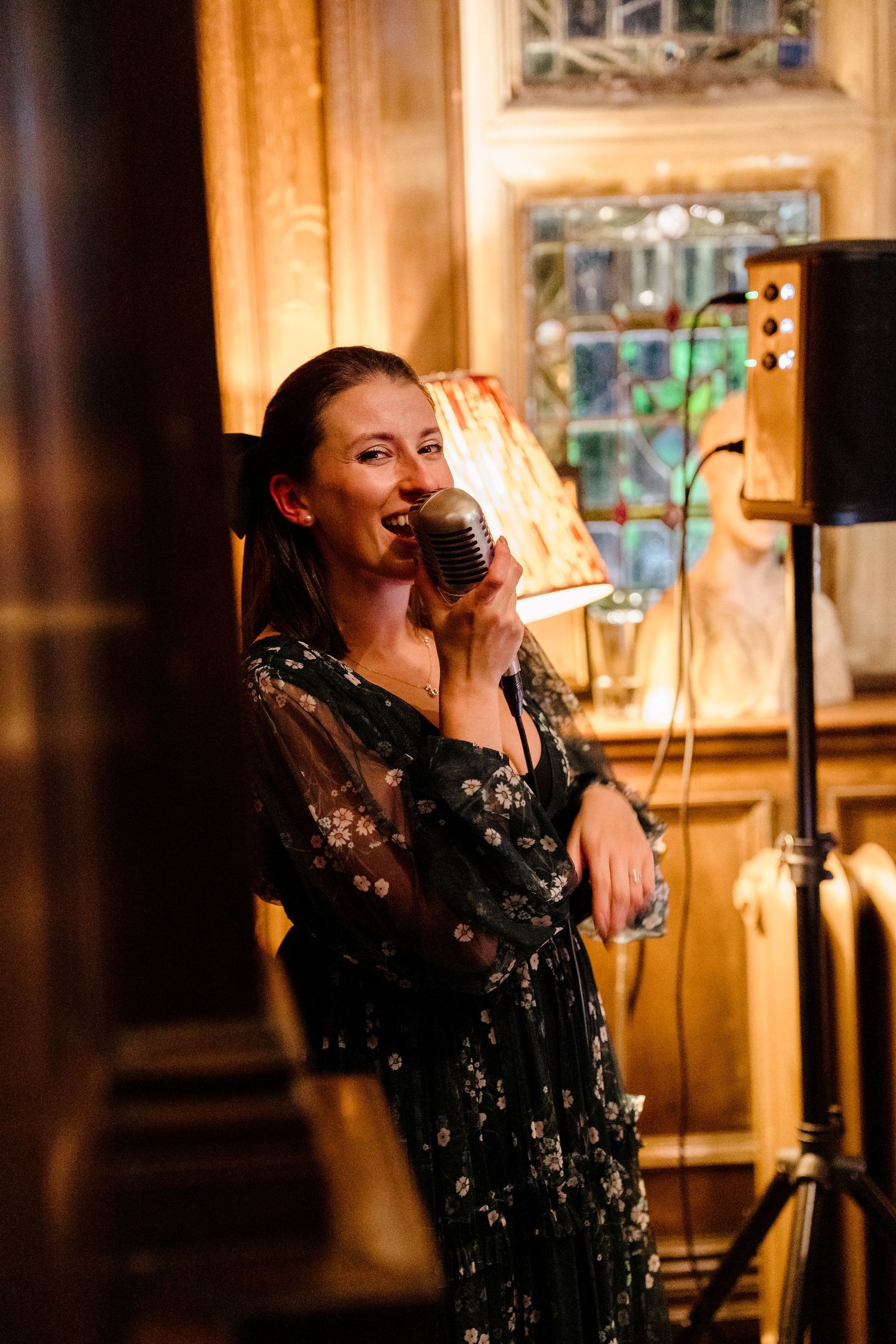 Woman in floral dress singing into vintage microphone in warmly lit elegant venue.