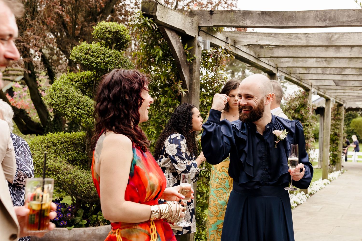 Guests in colorful attire laugh and chat at an outdoor wedding reception under a wooden pergola.