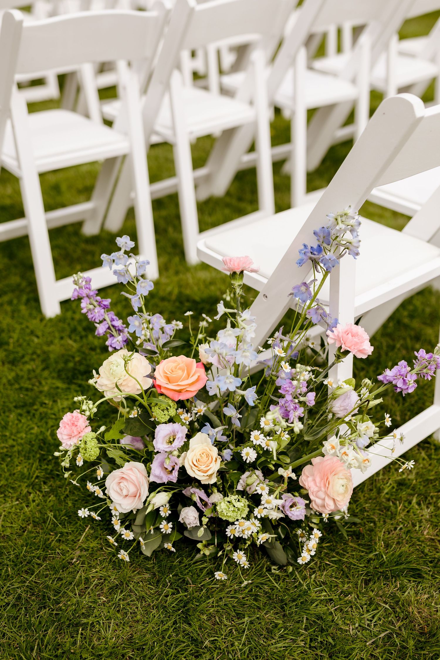 Colorful wedding aisle floral arrangement with pink roses and purple wildflowers beside white garden chairs.