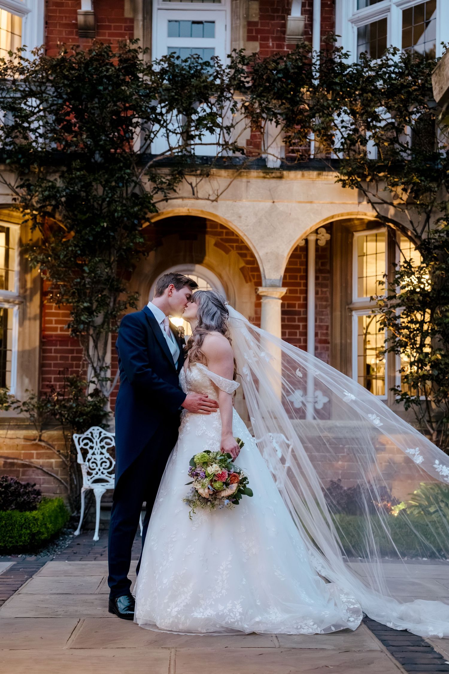 Bride and groom share a romantic kiss outside a brick manor, her veil flowing in the wind, holding a colorful bouquet.