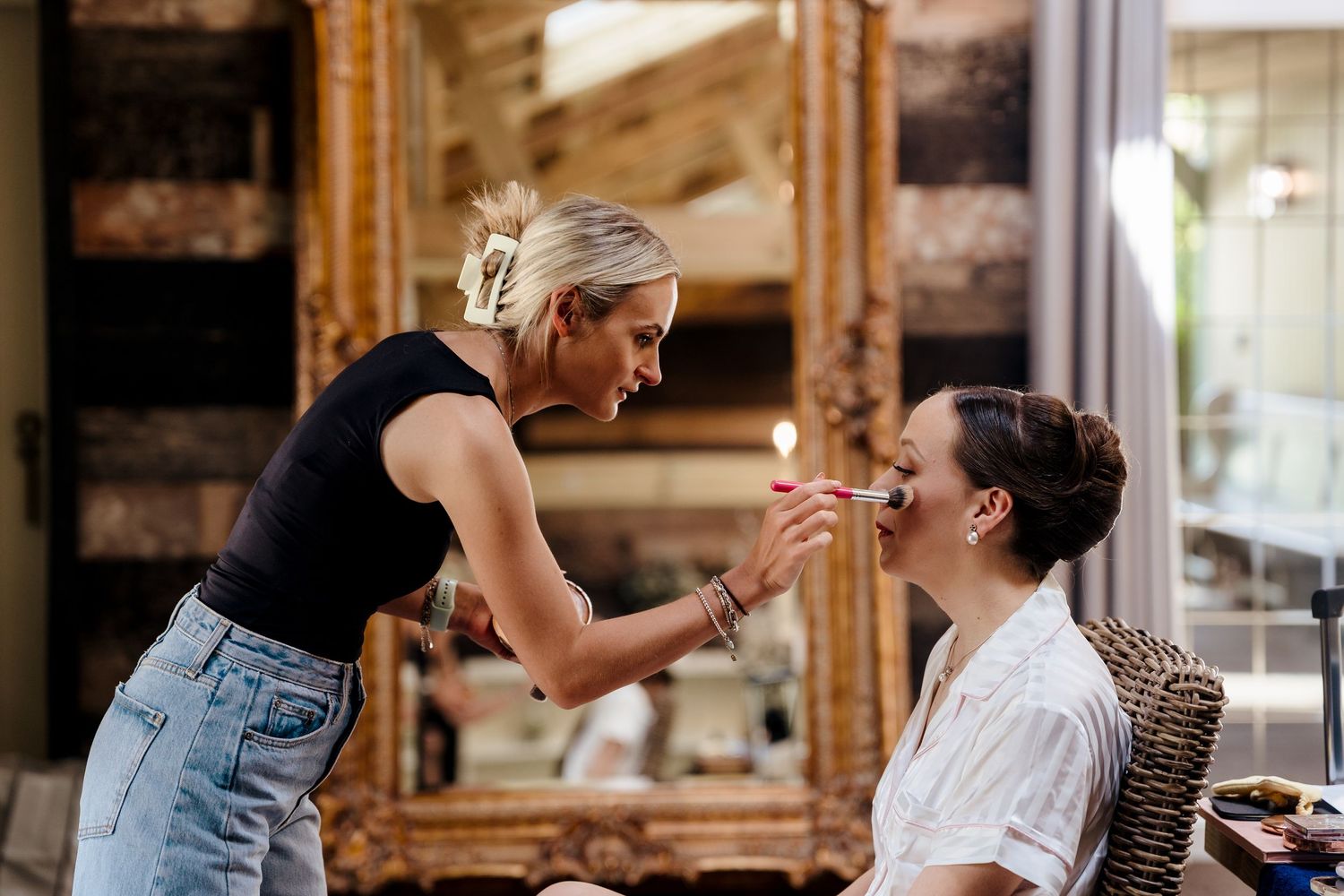 Professional makeup artist applying makeup to a seated client in front of an ornate gold mirror.
