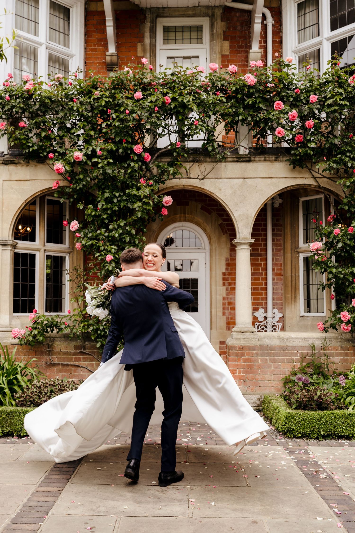 Groom lifting bride in white gown outside rose-covered brick manor, sharing joyful wedding moment.