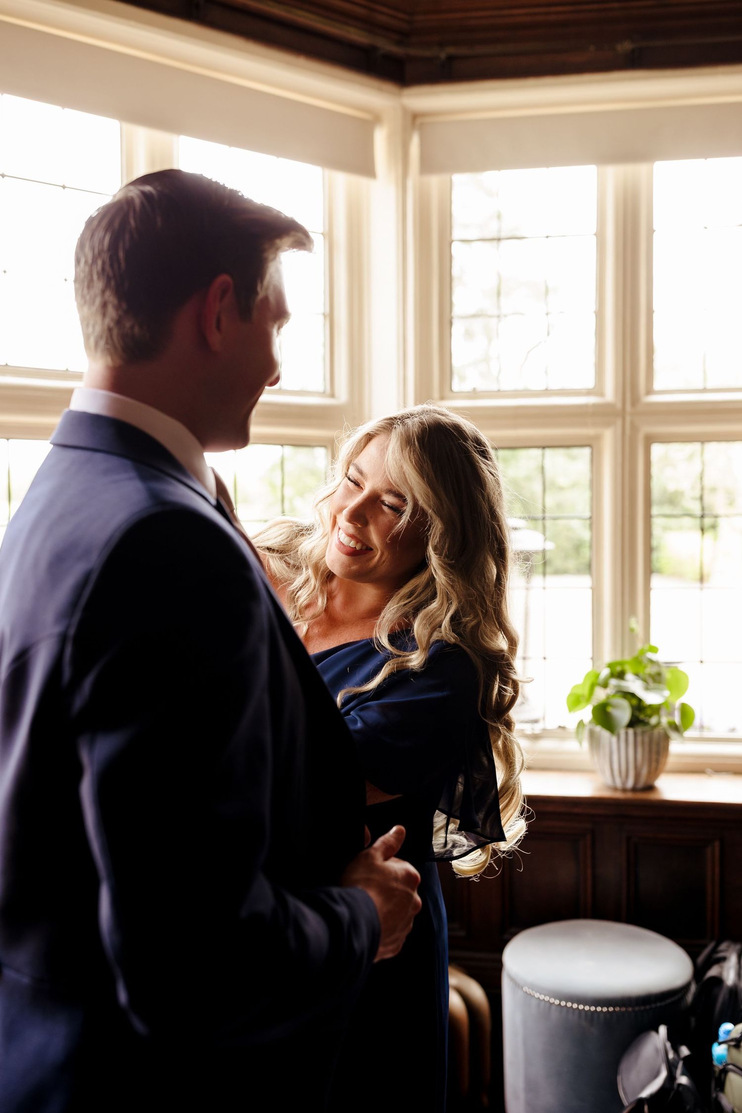 Groom in navy suit having tie adjusted by woman in blue dress near sunlit bay window.