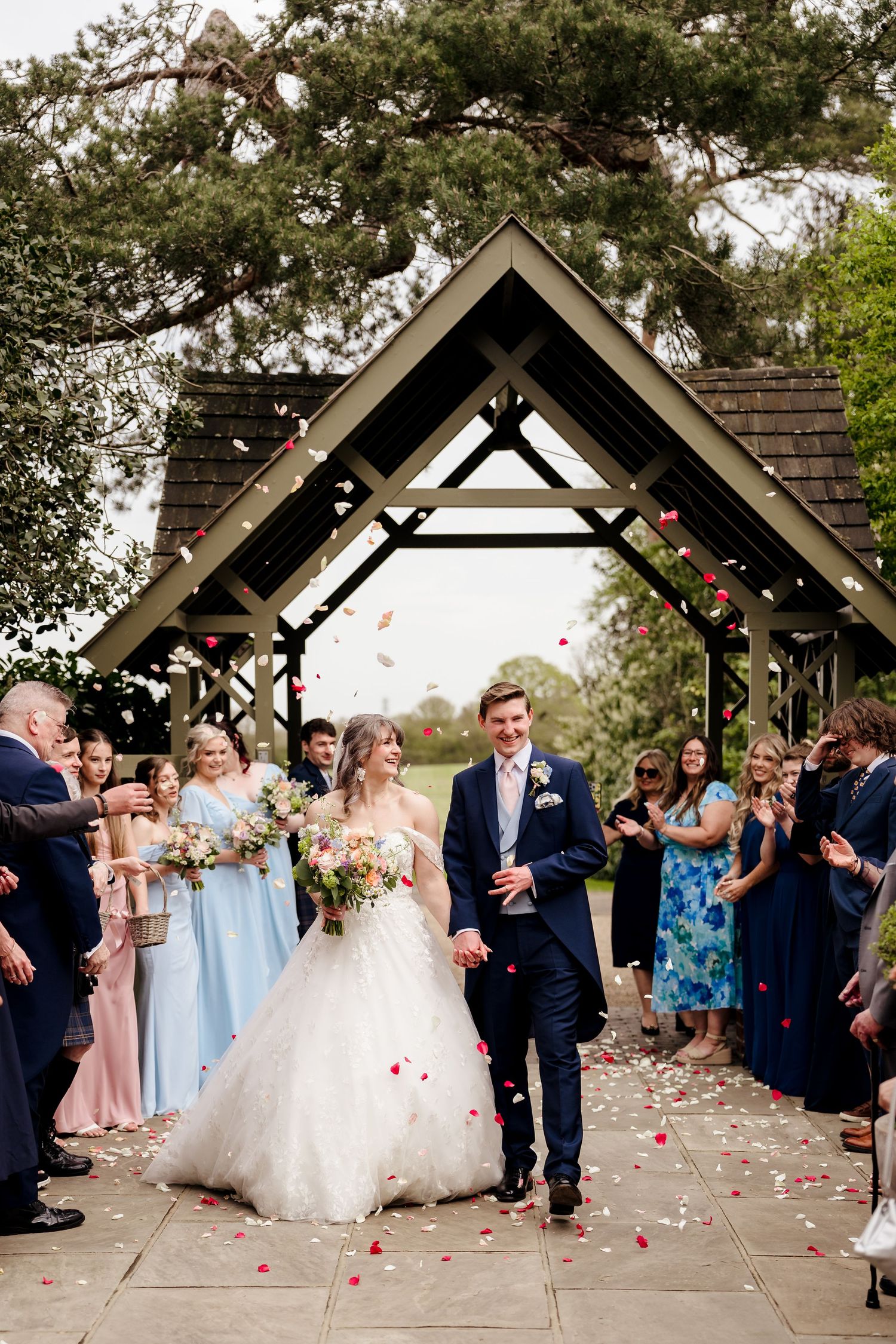 Bride and groom walk through a confetti shower under a wooden arch with guests celebrating.