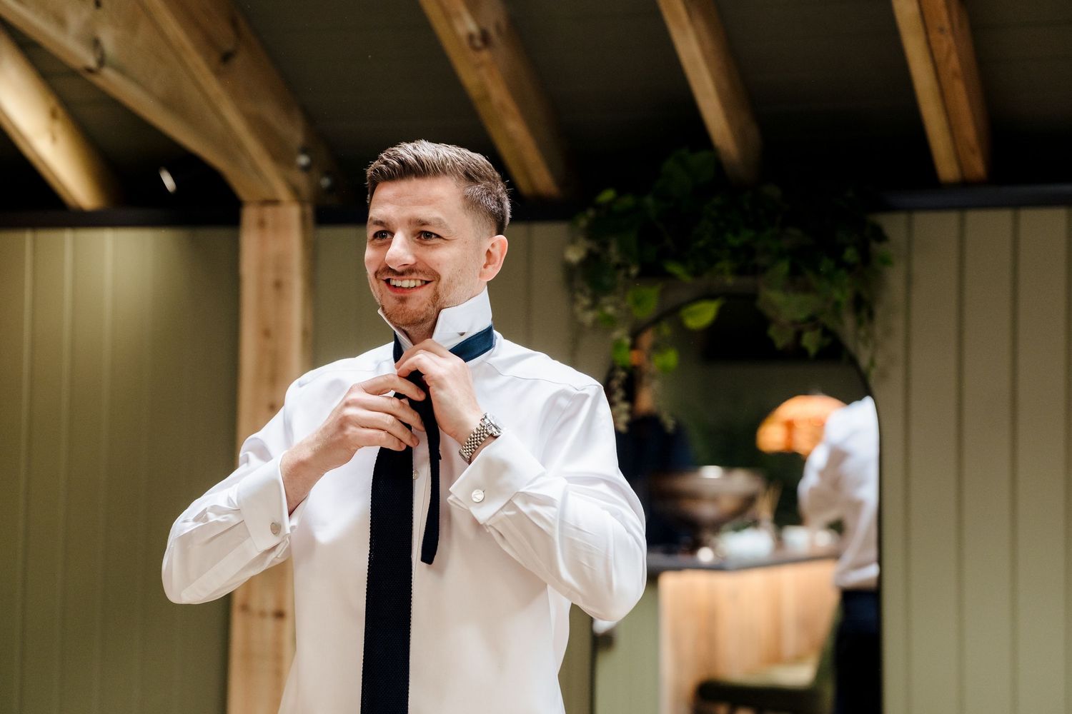 Smiling groom in white shirt adjusting his navy blue tie while getting ready for his wedding day.