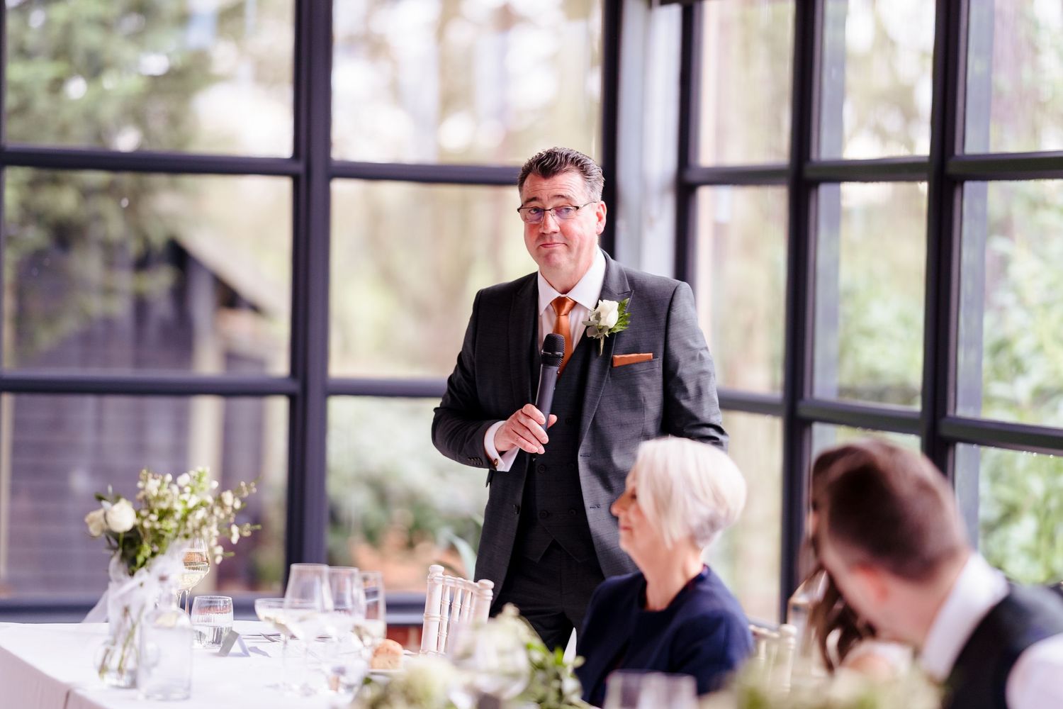Groom in dark suit delivers wedding speech to seated guests in bright windowed reception venue.