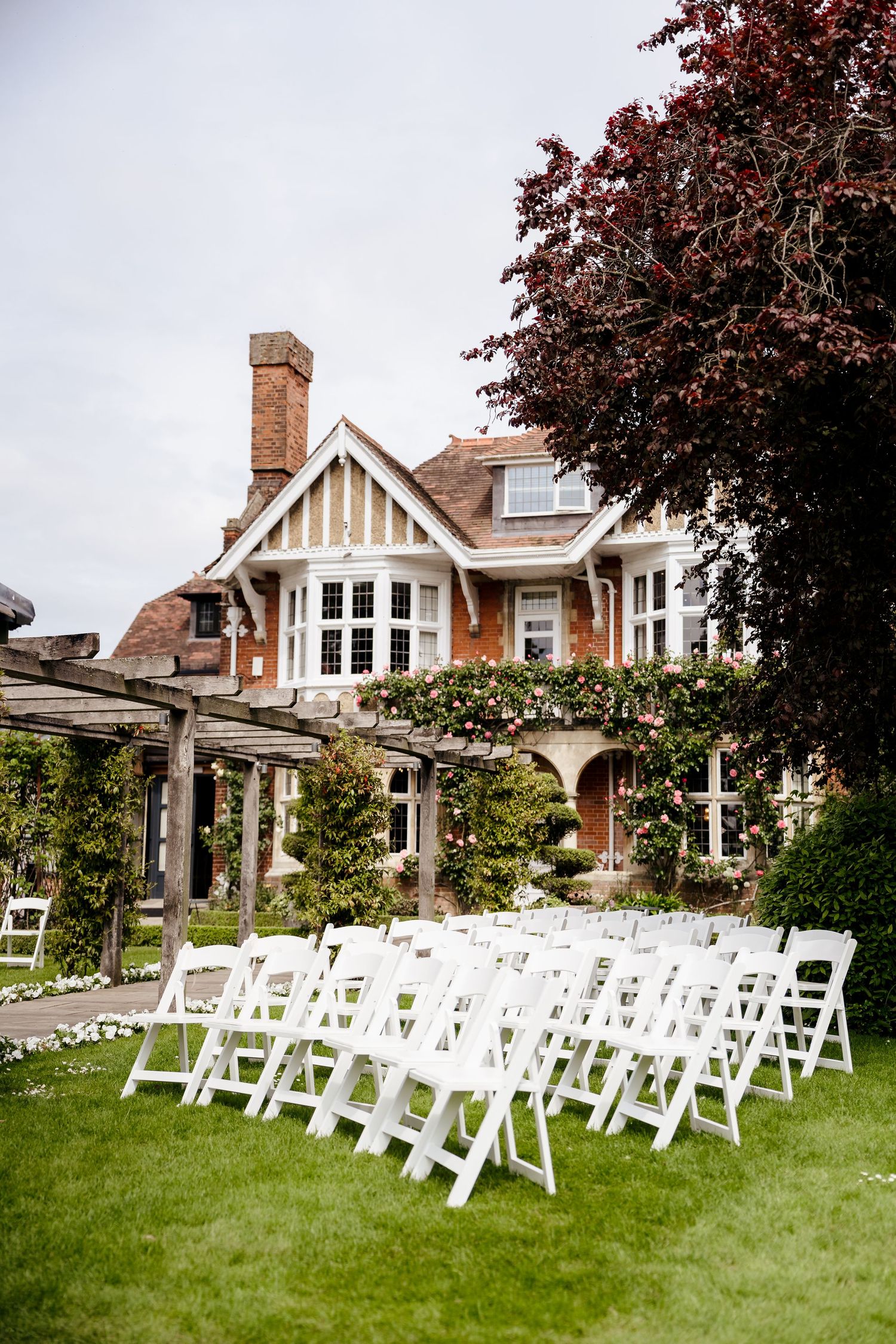 White folding chairs arranged on green lawn before Tudor-style brick manor house for outdoor wedding ceremony.