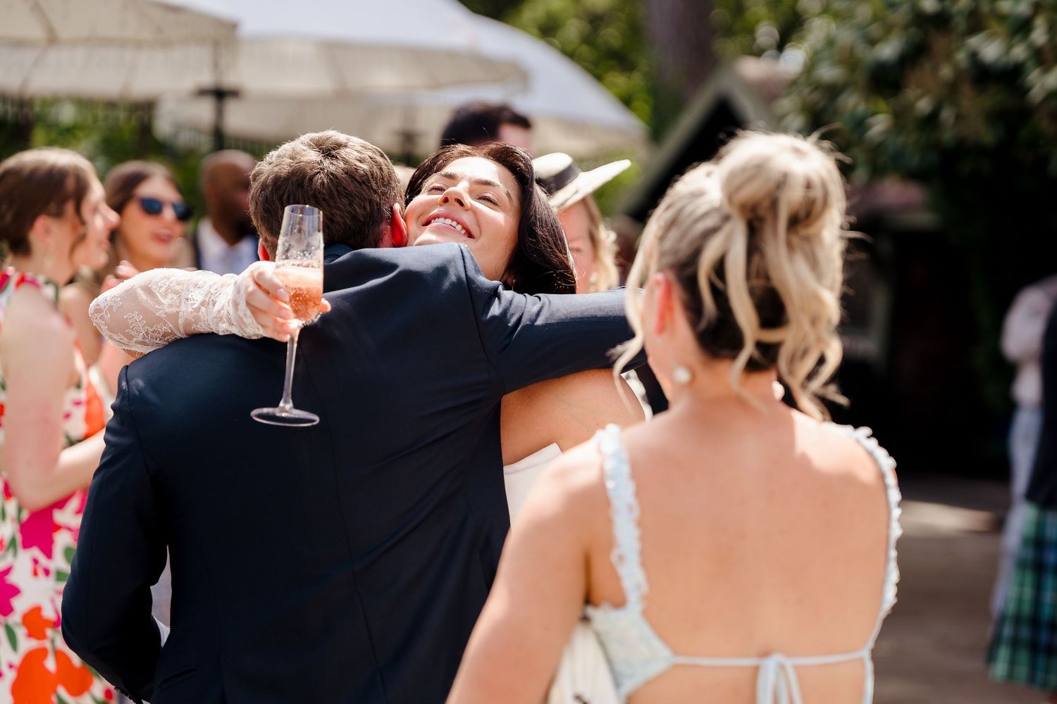 Guests embracing and celebrating at an outdoor wedding reception, holding champagne glasses in sunny venue.