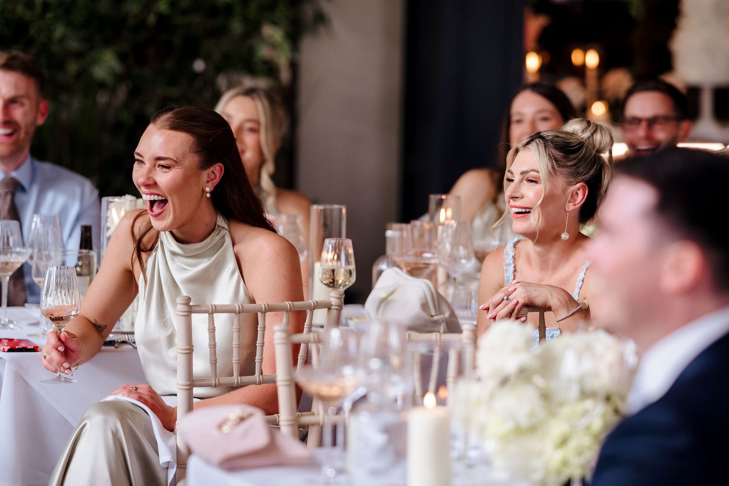 Guests laughing and smiling at a wedding reception dinner, dressed in elegant attire at a beautifully set table.