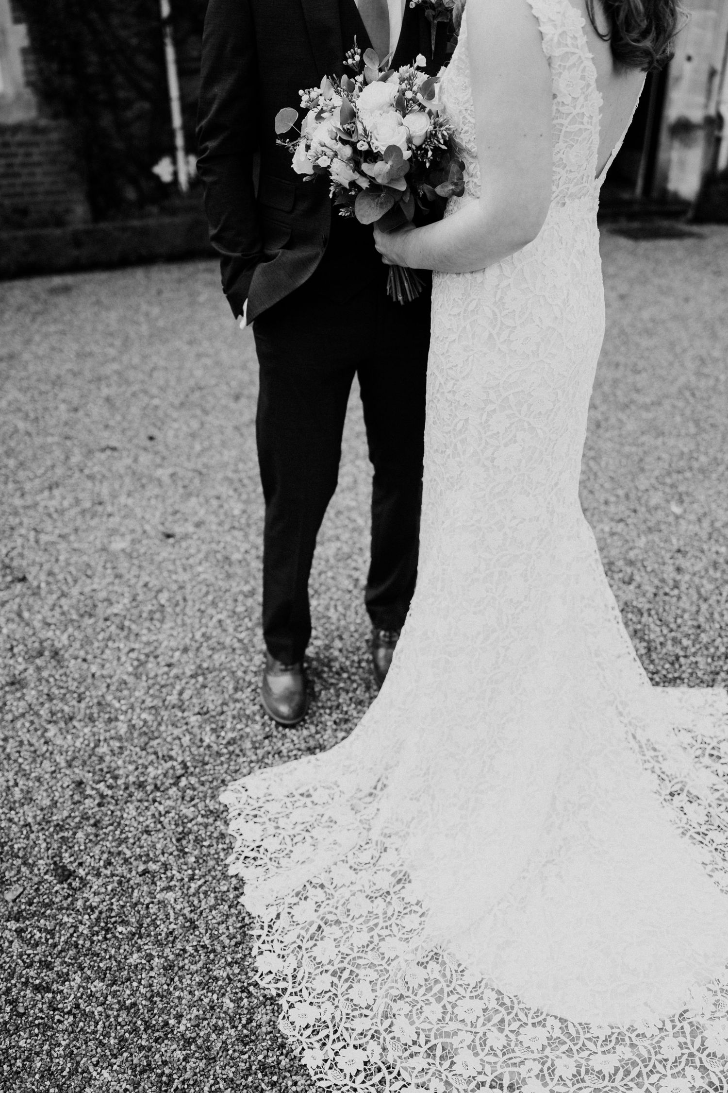 Black and white photo of bride and groom with lace train spread on gravel path.
