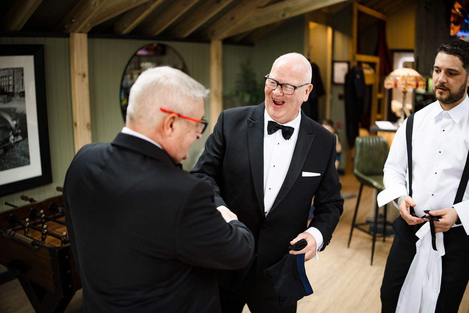 Men in formal tuxedos laughing and socializing at an elegant indoor wedding reception event.