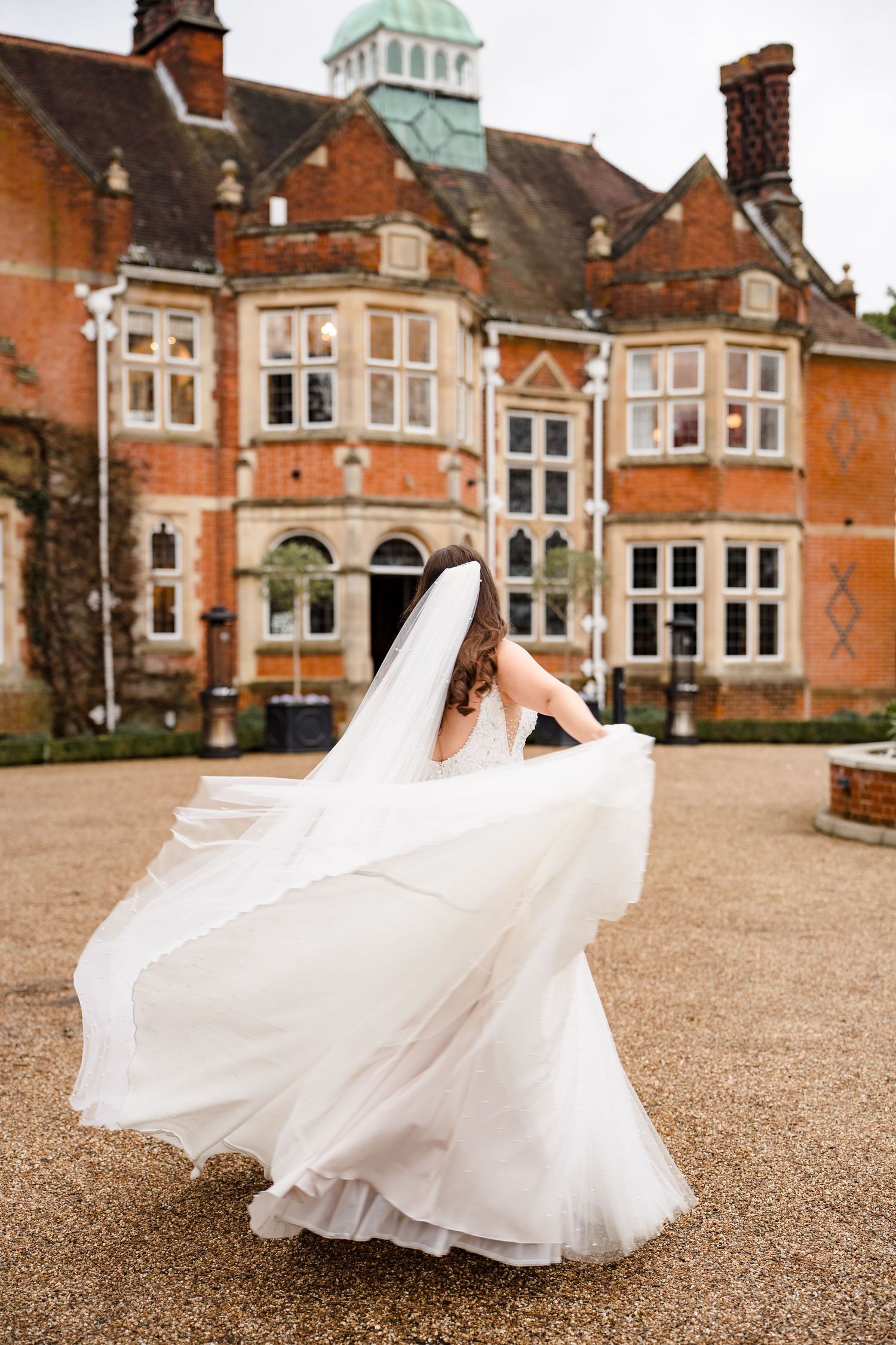 Bride twirls her flowing white wedding dress and veil outside a grand red brick manor house.