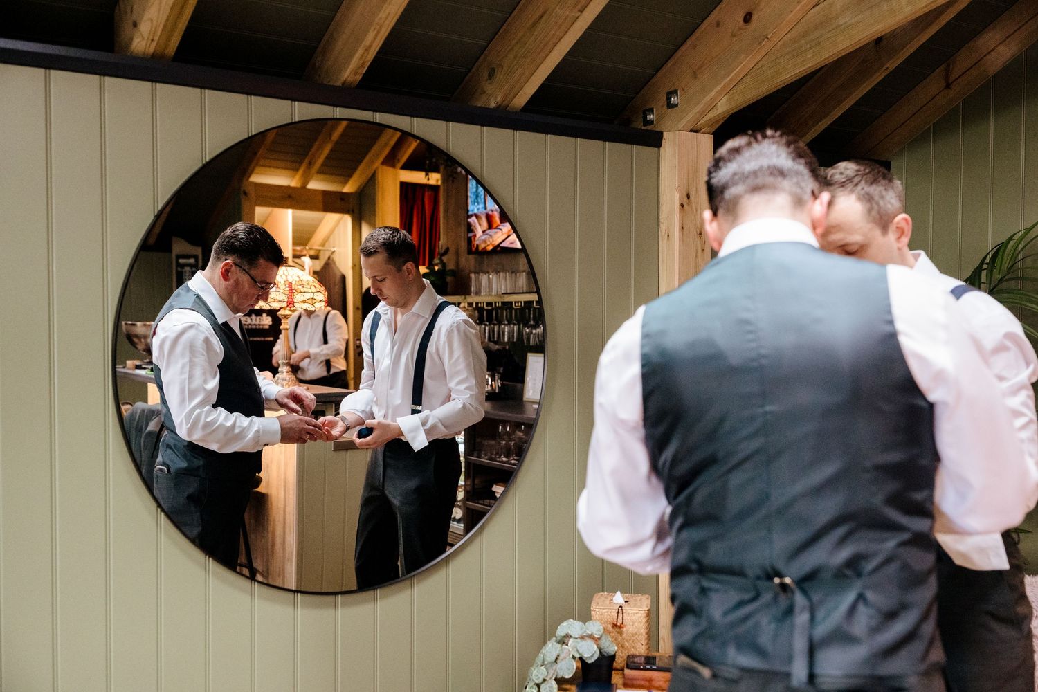 Groomsmen in white shirts and suspenders prepare for a wedding, reflected in a round mirror.