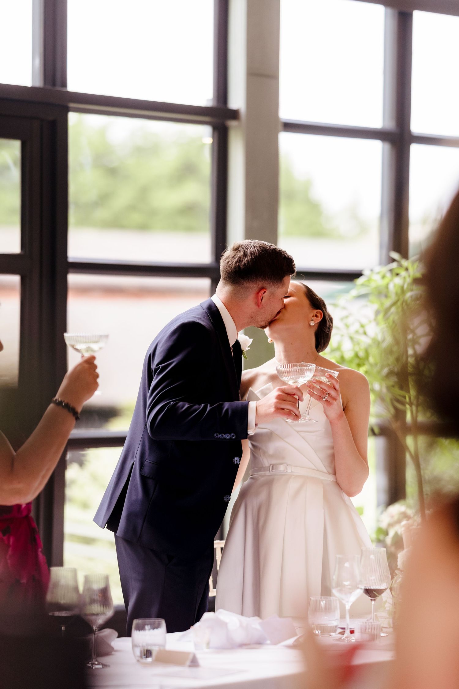 Bride and groom sharing a kiss while holding champagne glasses during wedding reception indoors.