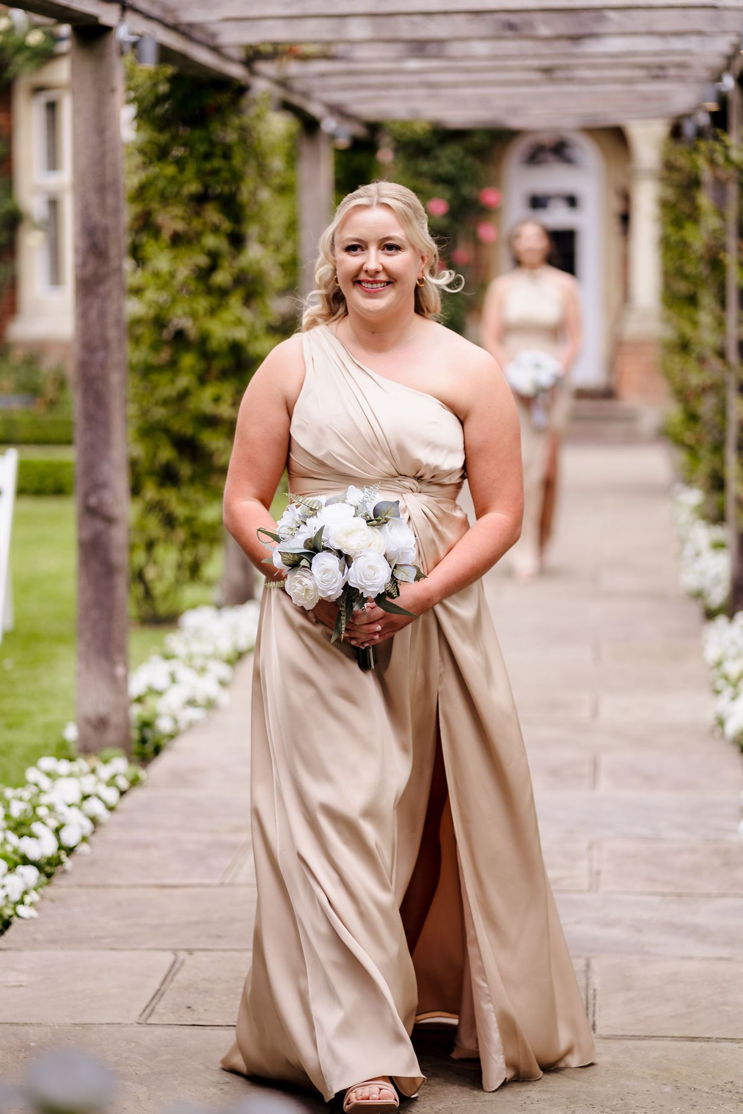 Bridesmaid in gold one-shoulder gown walks down garden path holding white bouquet at elegant wedding venue.