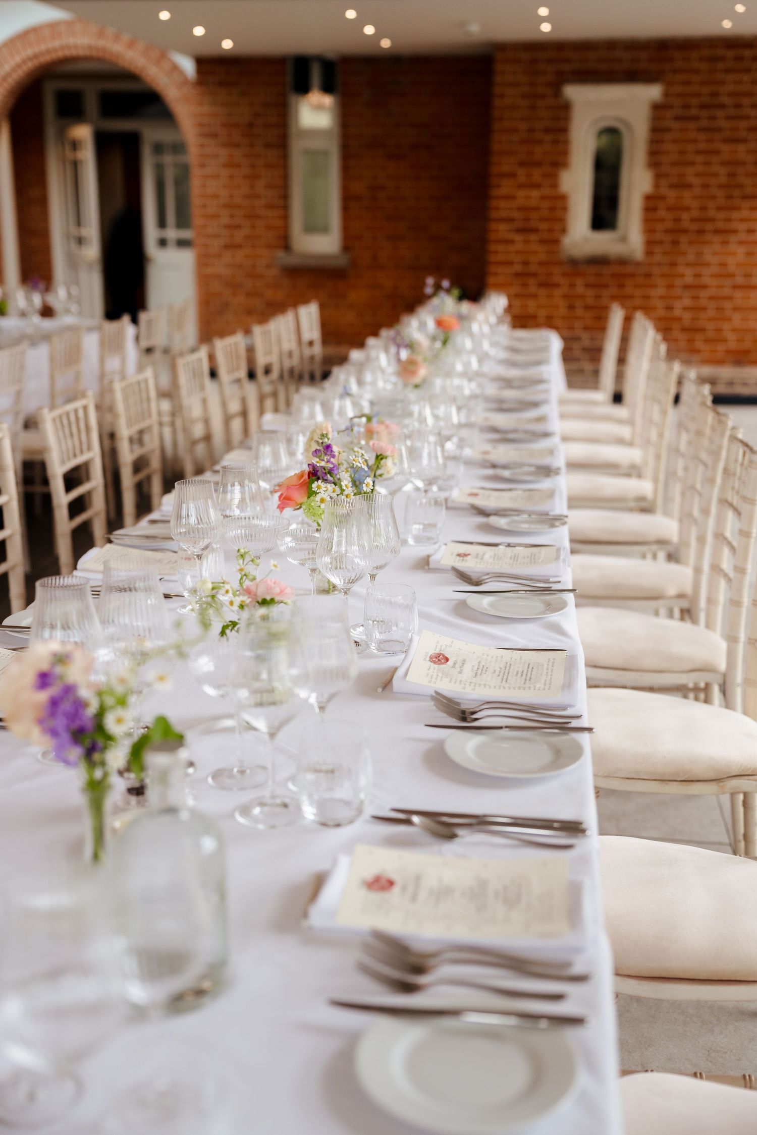 Elegant wedding reception long table with white linens, floral centerpieces, glassware, and place settings in a brick venue.