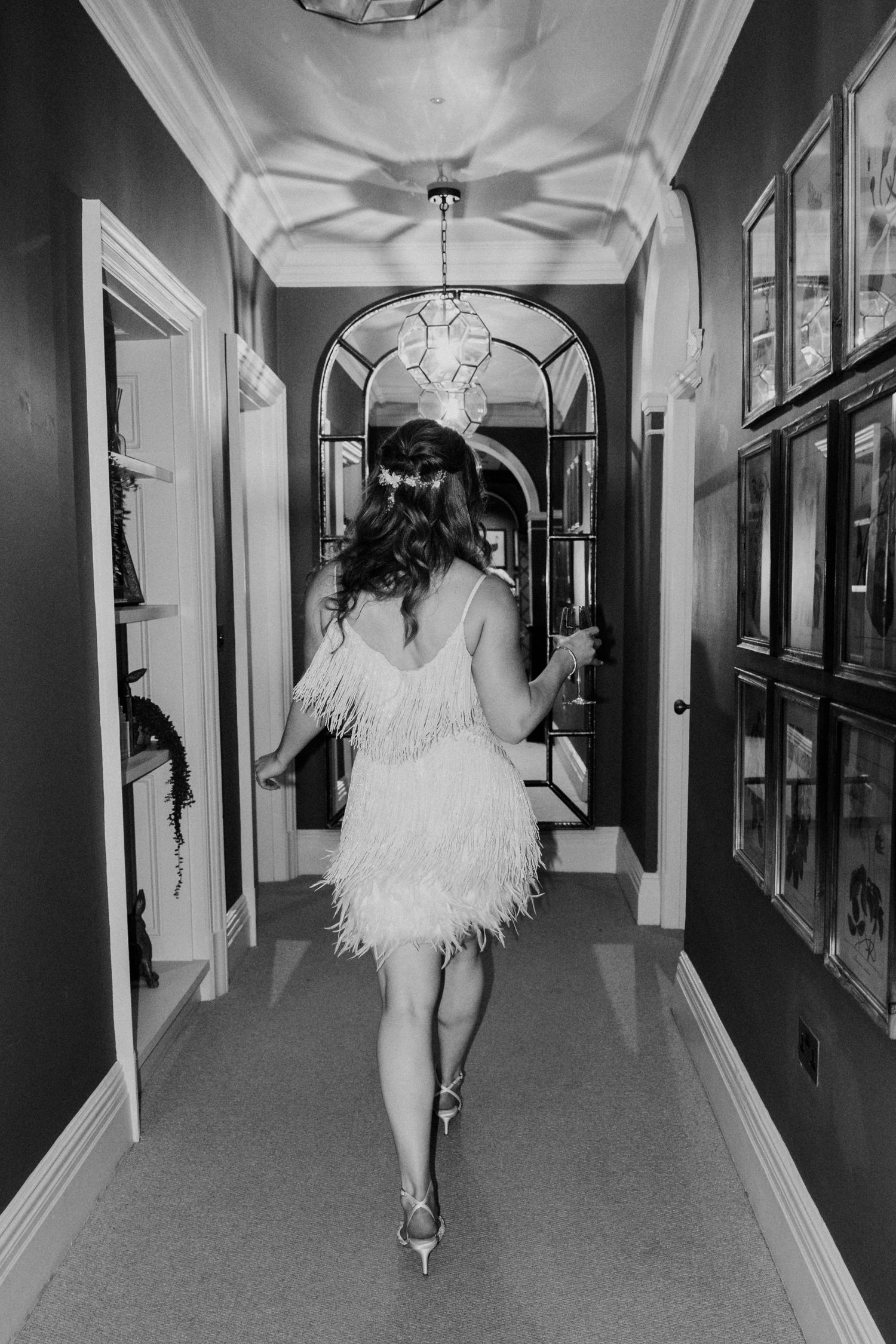Black and white photo of bride in feathered mini dress walking down elegant hotel corridor.