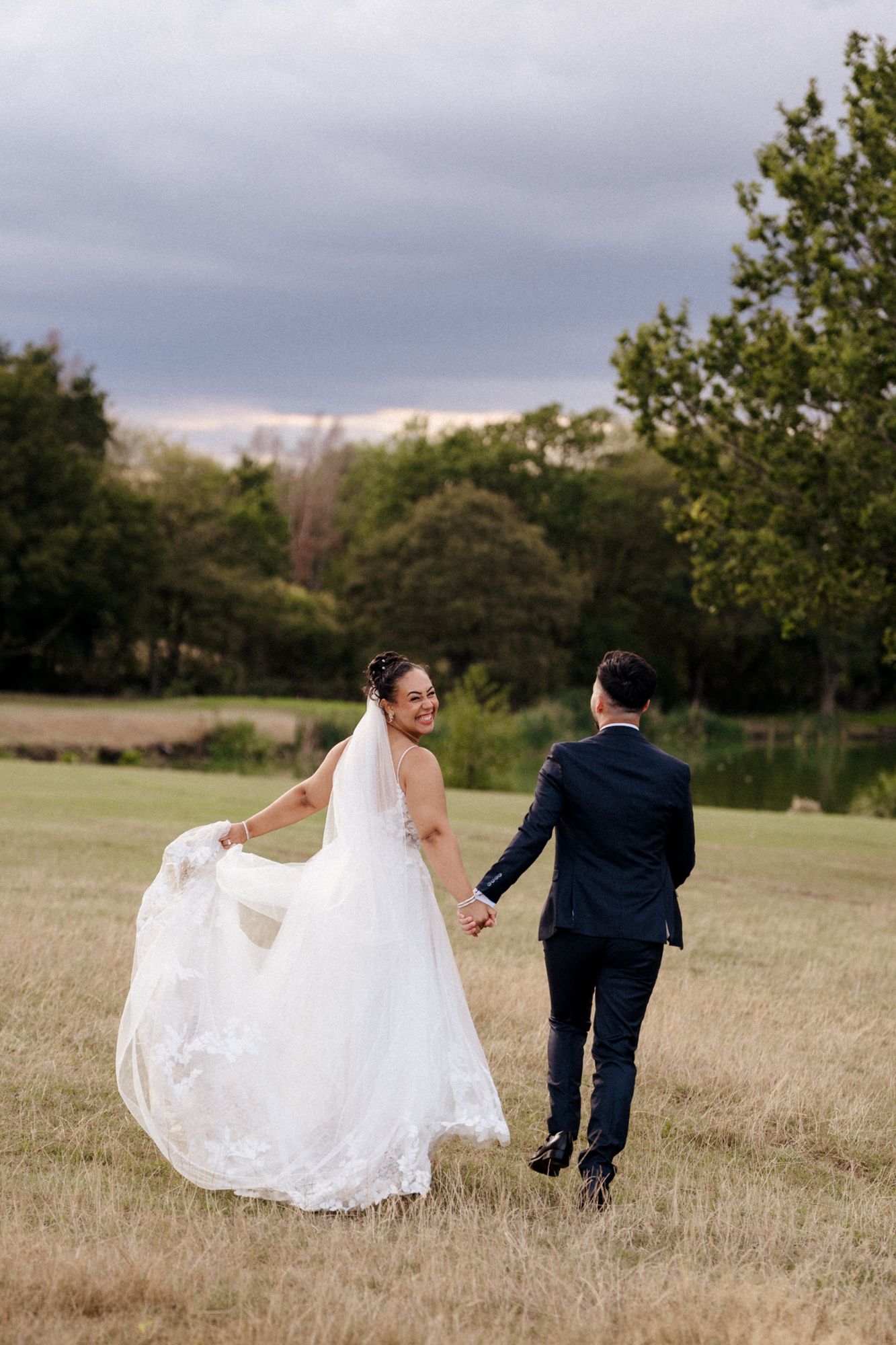 Bride in white lace wedding dress and groom in navy suit walking hand in hand across a field with trees in background.