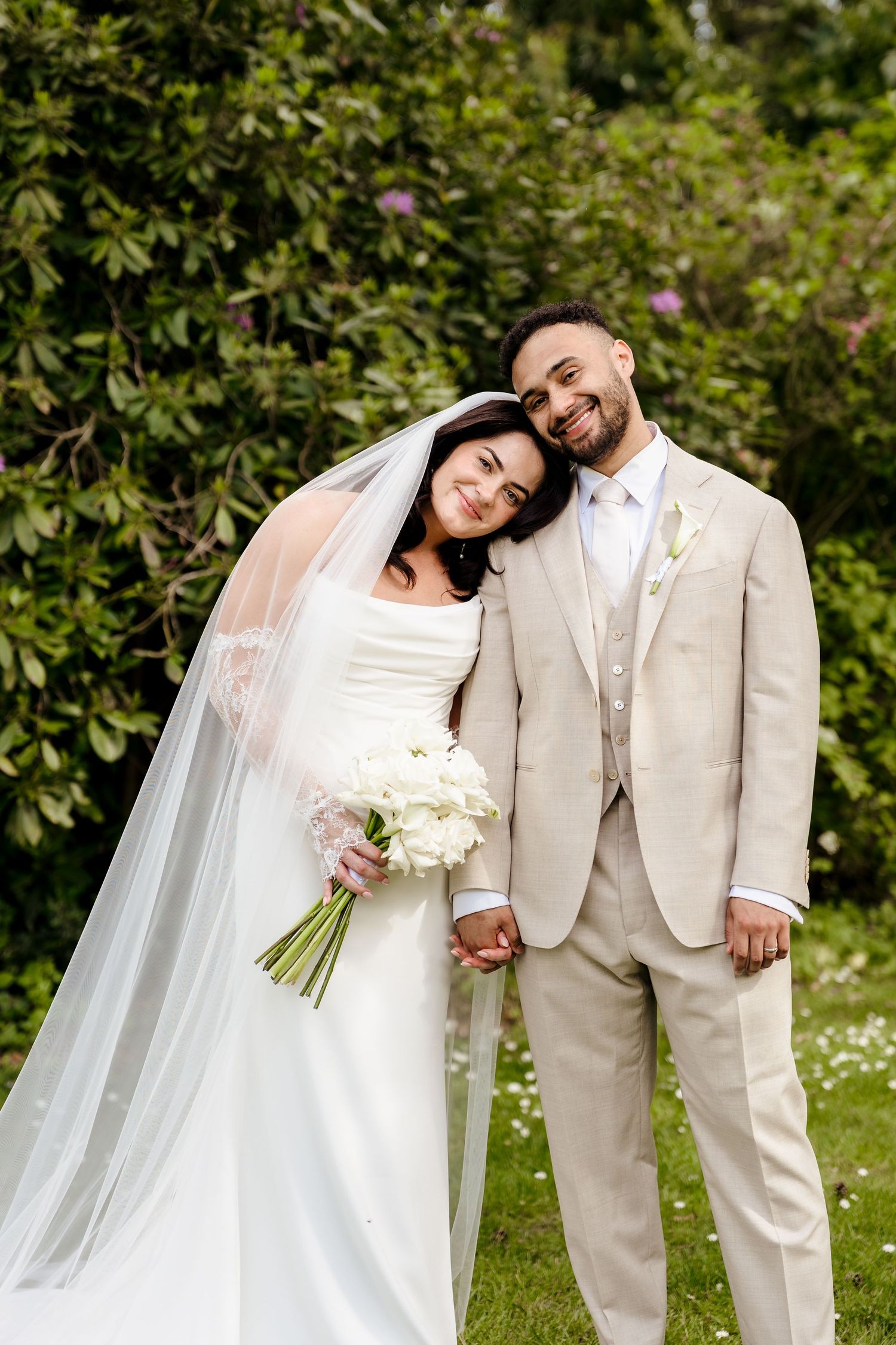 Bride and groom posing together in garden, bride holding white bouquet in elegant wedding attire.