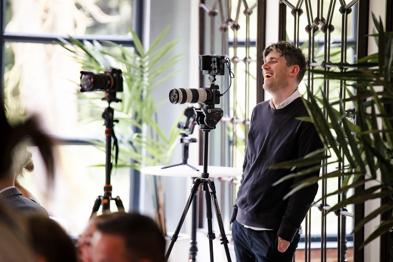 Man laughing near camera tripods with professional photography equipment indoors, surrounded by plants and natural light.