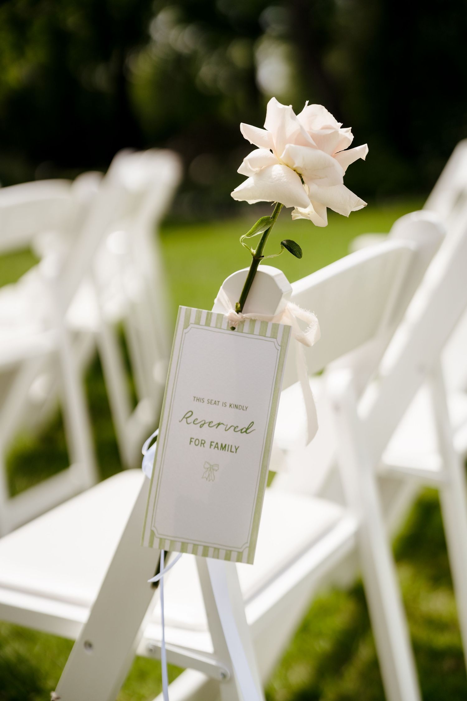 White folding chairs with a reserved for family sign and single white rose at an outdoor wedding ceremony.