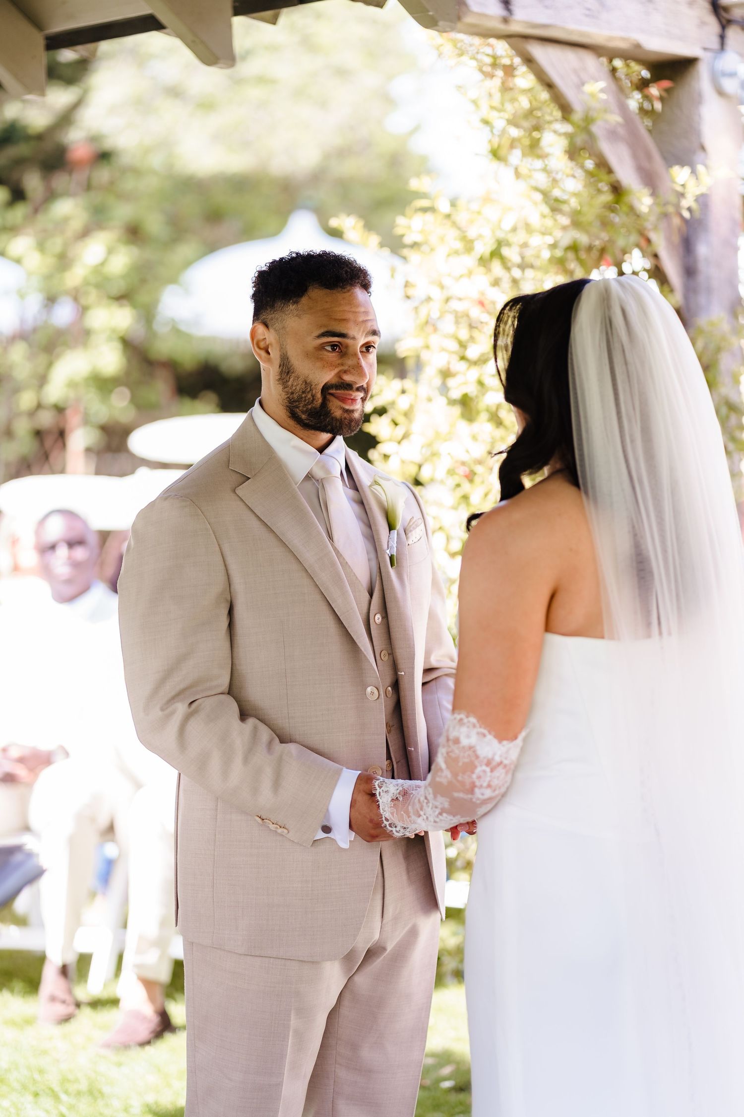Groom in beige suit smiles at bride in white lace wedding dress during outdoor ceremony.
