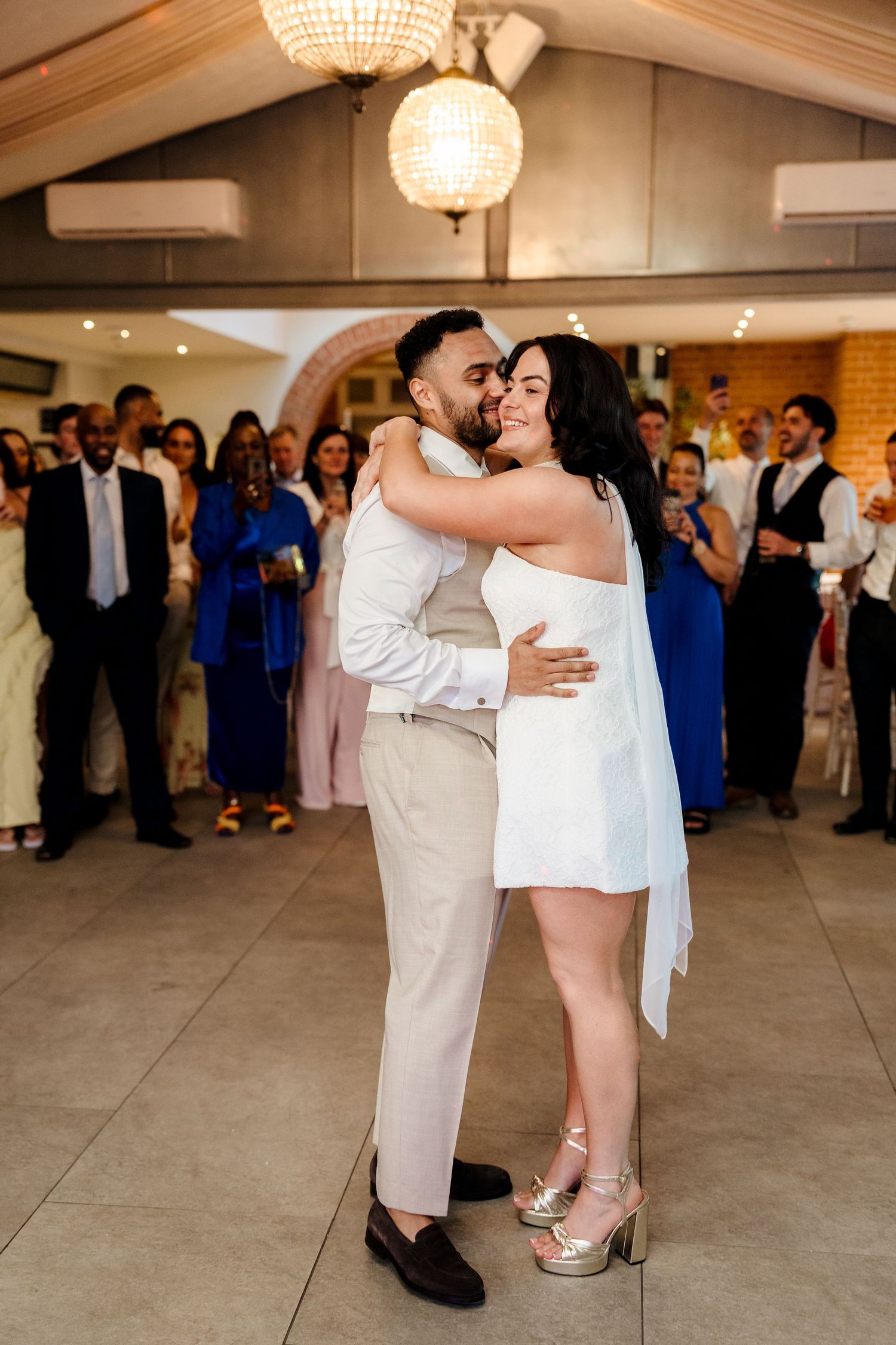 Bride in short white dress and groom share first dance embrace under chandelier at wedding reception.