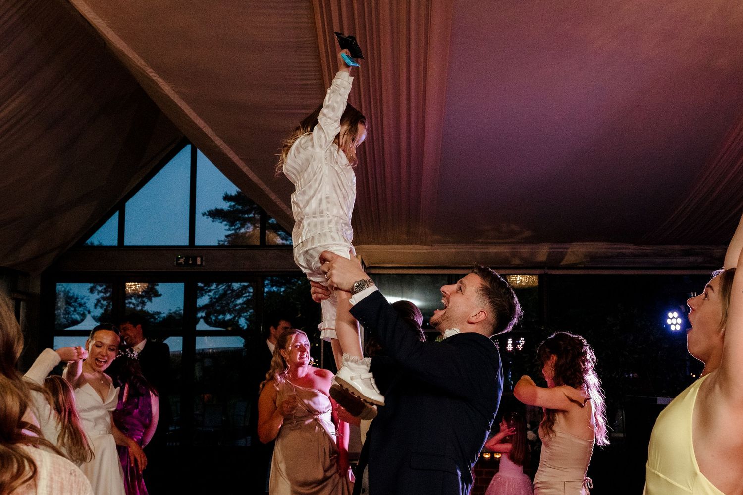 Groom lifts bride above crowd of dancing wedding guests during energetic evening reception celebration under tent.