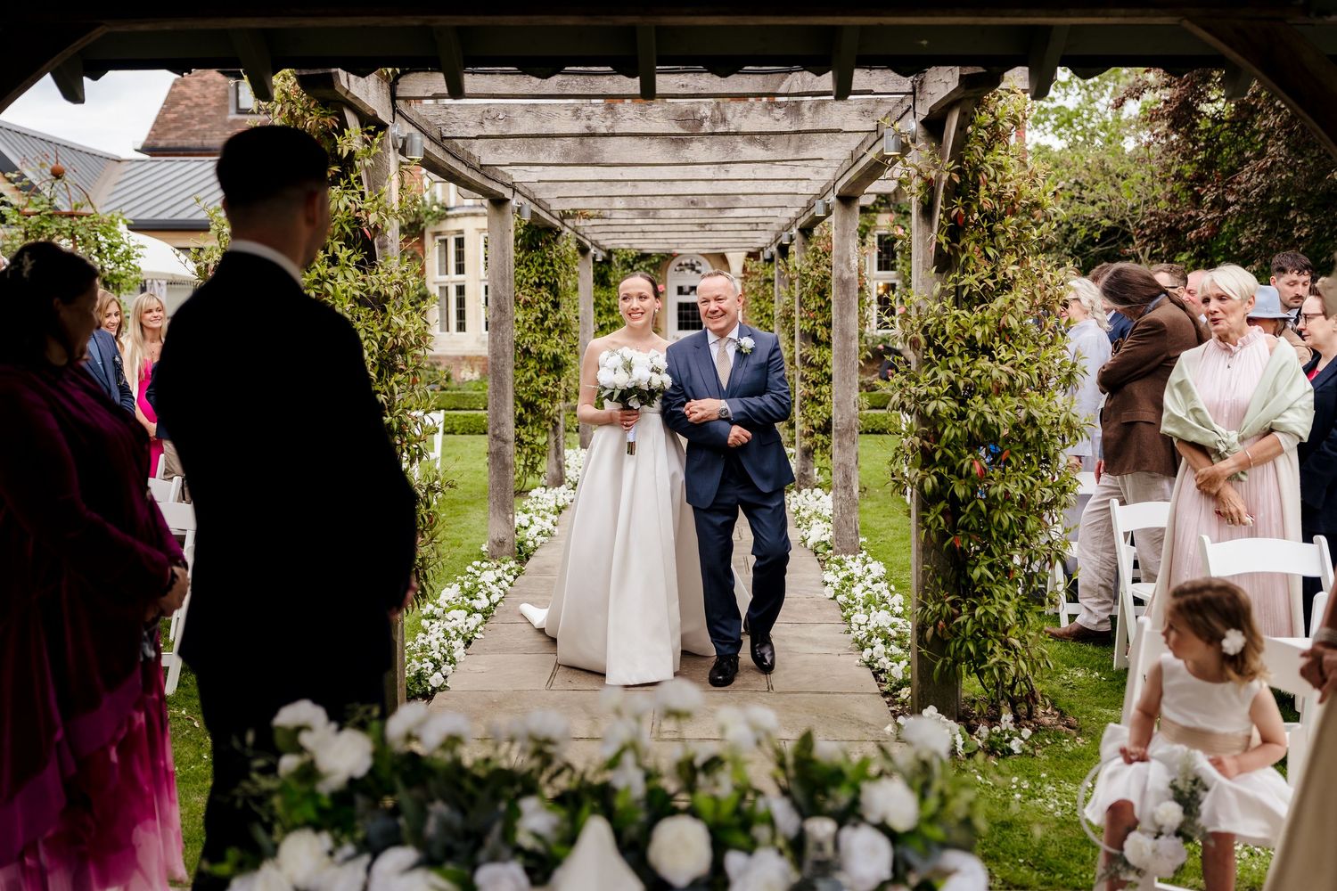 Bride walks down outdoor aisle under floral pergola with wedding guests lining the path.