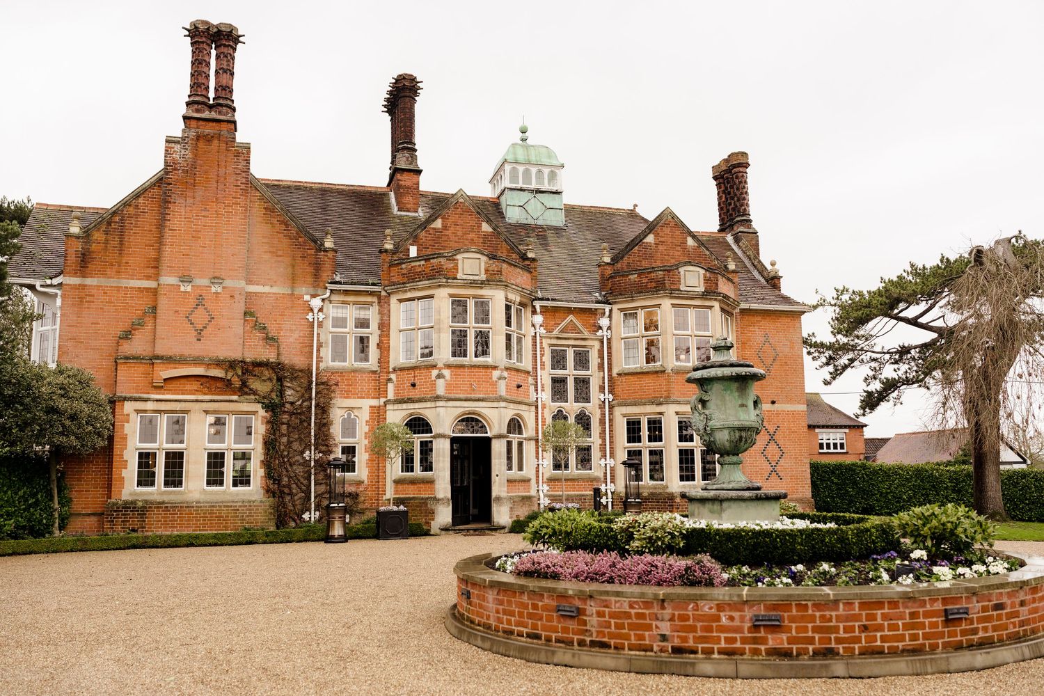 Grand red brick English manor house wedding venue with copper dome, ornate chimneys, and circular fountain garden.
