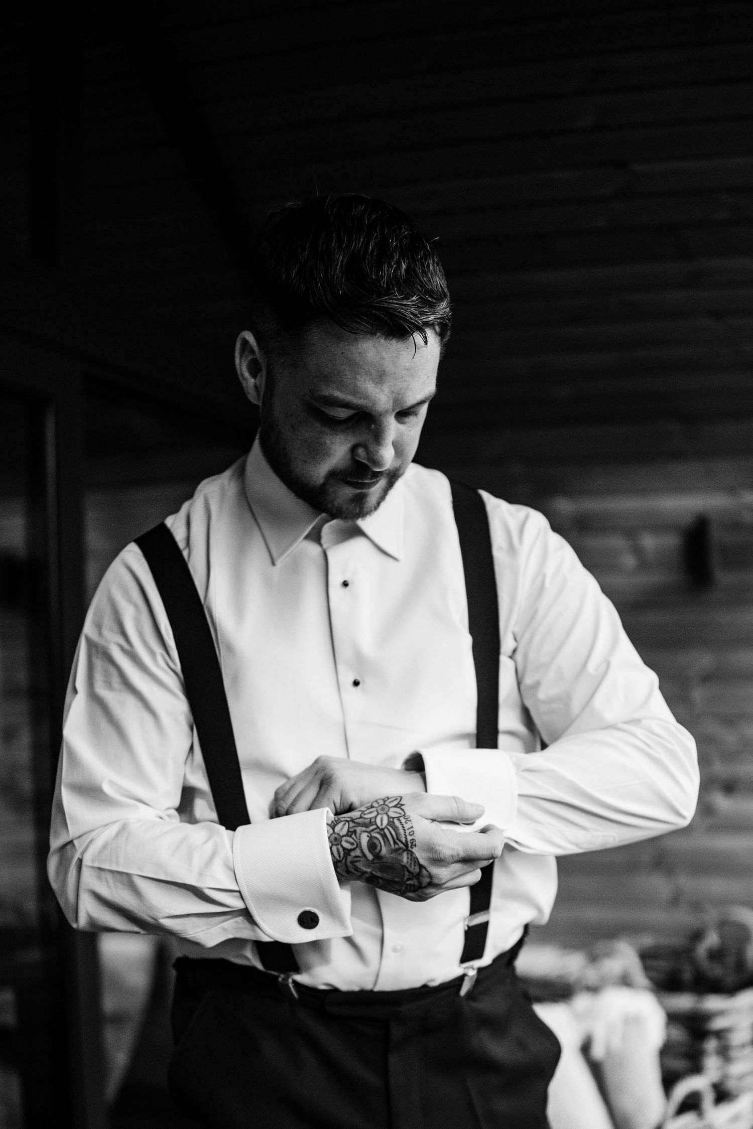 Groom in white dress shirt and suspenders adjusting his cufflinks, with a tattoo visible on his hand.