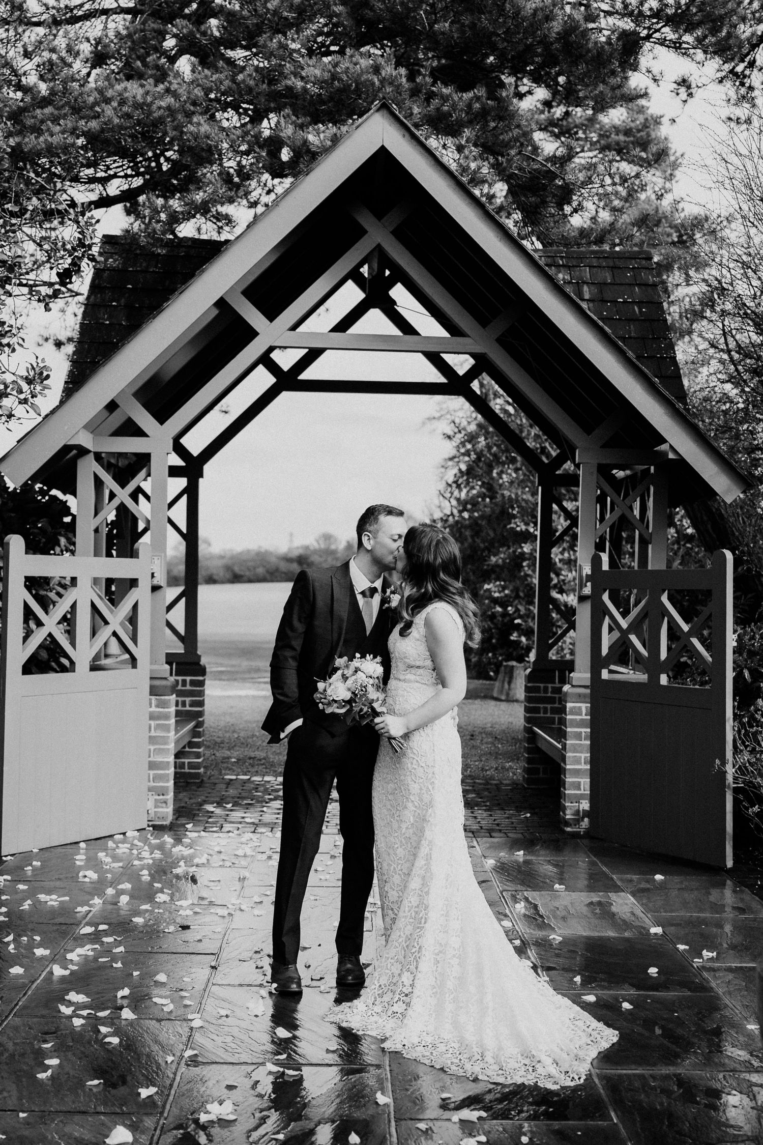 Black and white photo of bride and groom kissing under wooden arch gateway on rainy wedding day.