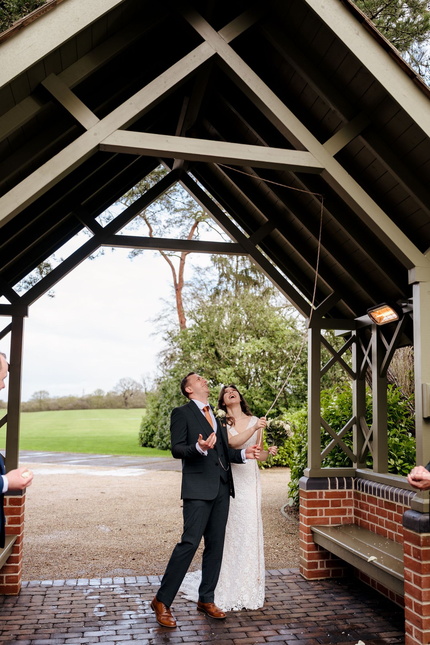 Bride and groom laugh joyfully under a wooden pergola as confetti falls at their wedding.