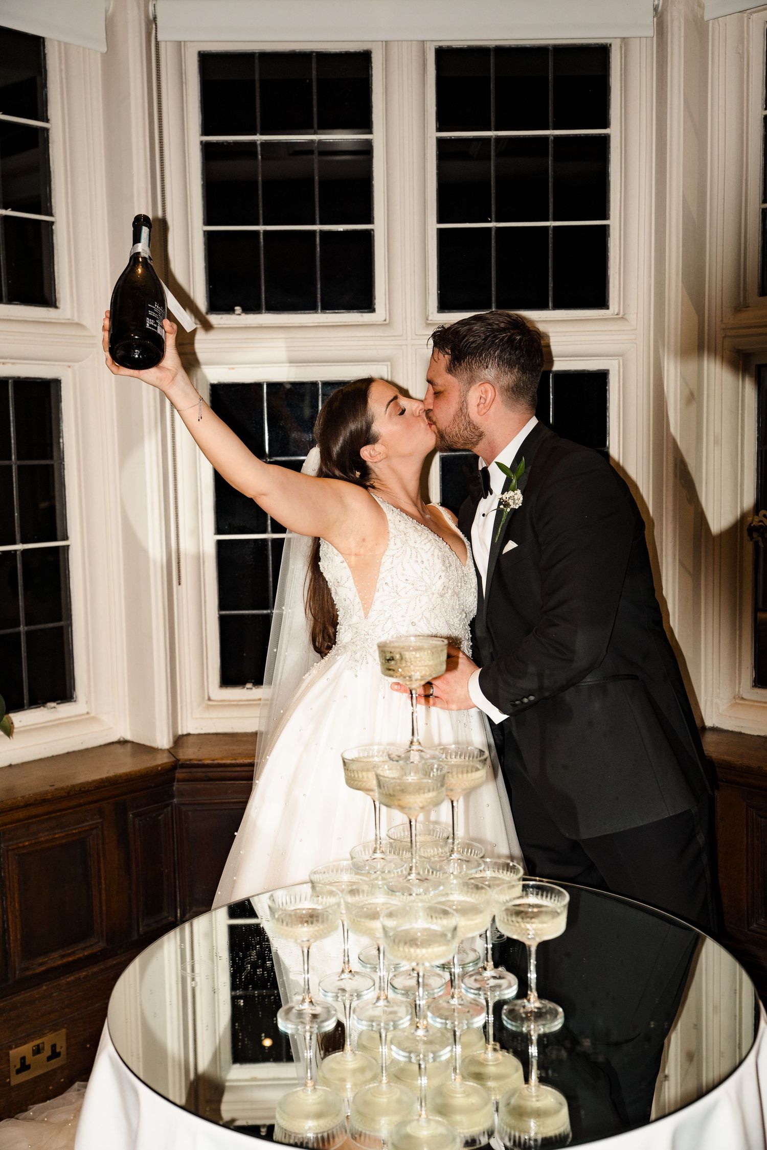 Bride and groom kissing beside champagne tower, holding bottle aloft at elegant wedding reception.