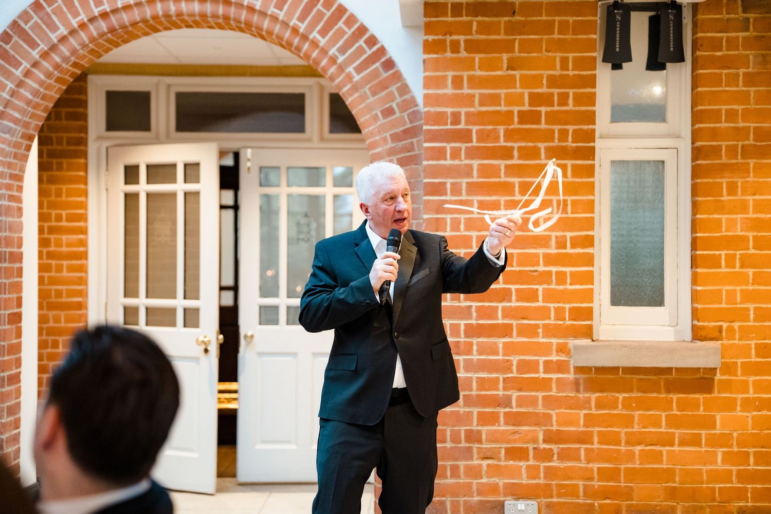 Older gentleman in dark suit speaking into microphone during outdoor wedding speech at brick venue.