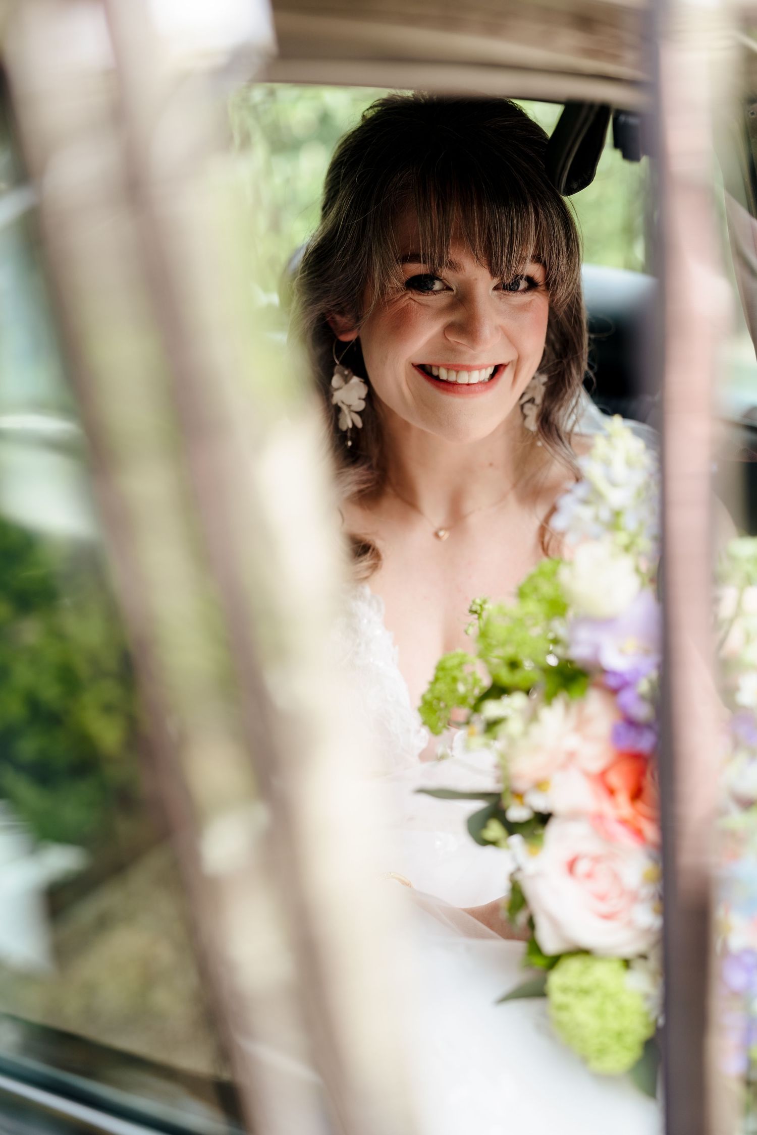 Radiant bride smiling through vintage car window holding colorful bridal bouquet of pink and green flowers.