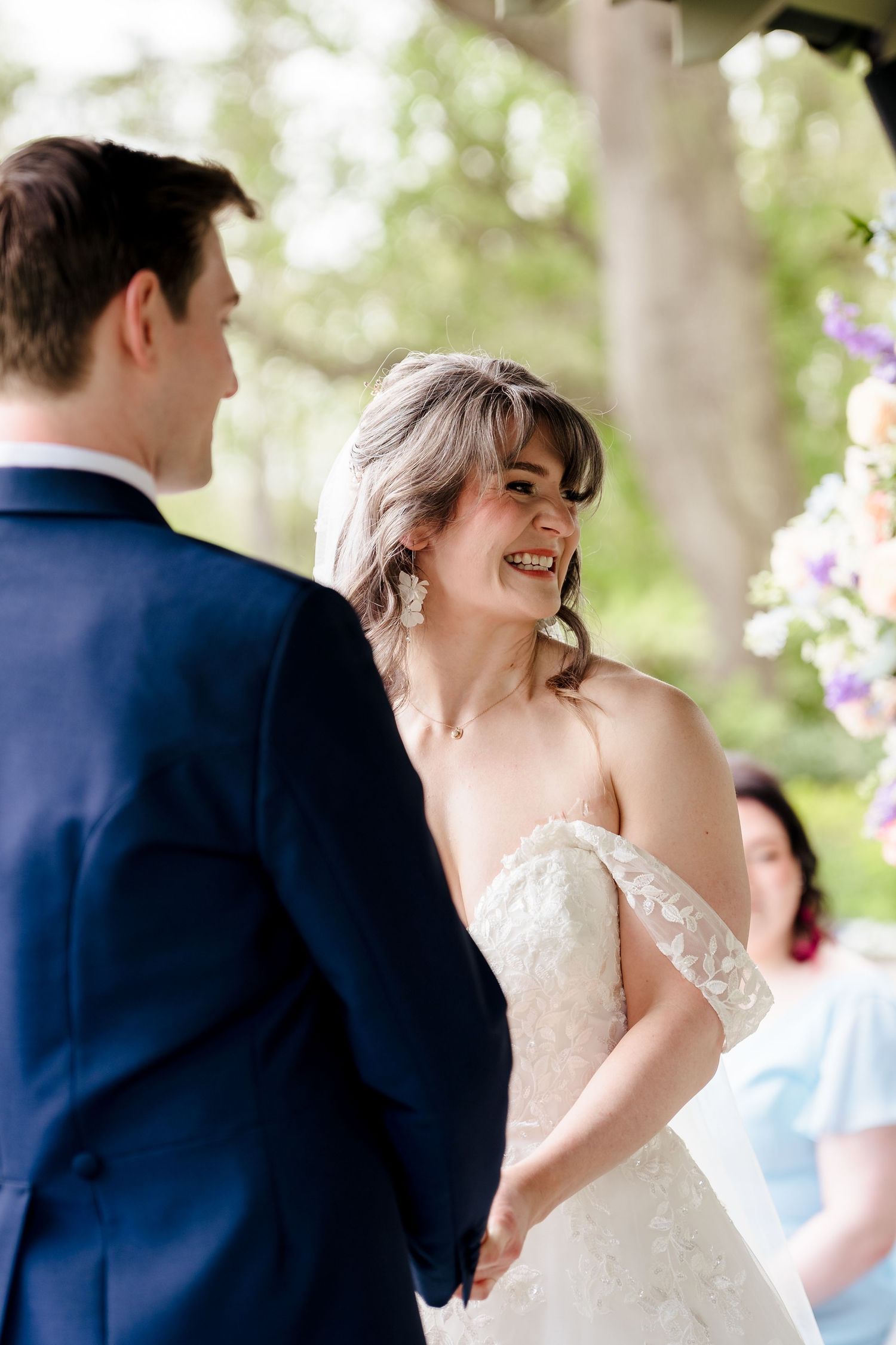 Smiling bride in off-shoulder lace gown holding hands with groom during outdoor ceremony.
