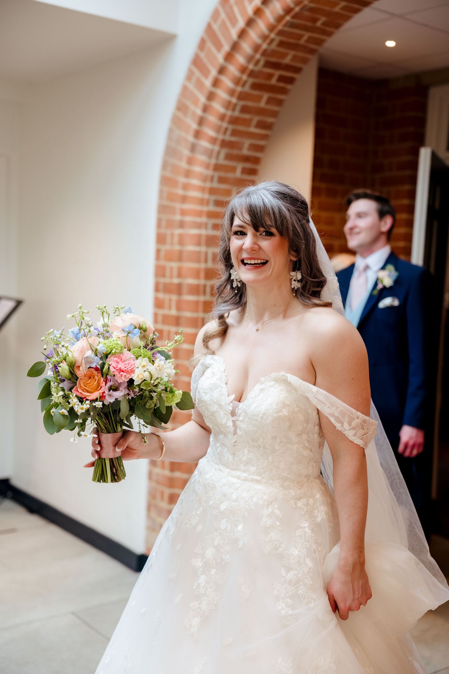 Radiant bride in lace off-shoulder gown holds colorful bouquet, laughing joyfully inside brick archway.