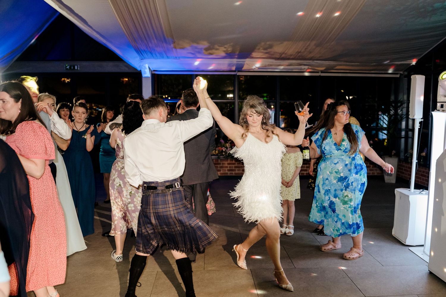 Wedding guests dancing energetically on the dance floor, including a man in a Scottish kilt at a reception party.