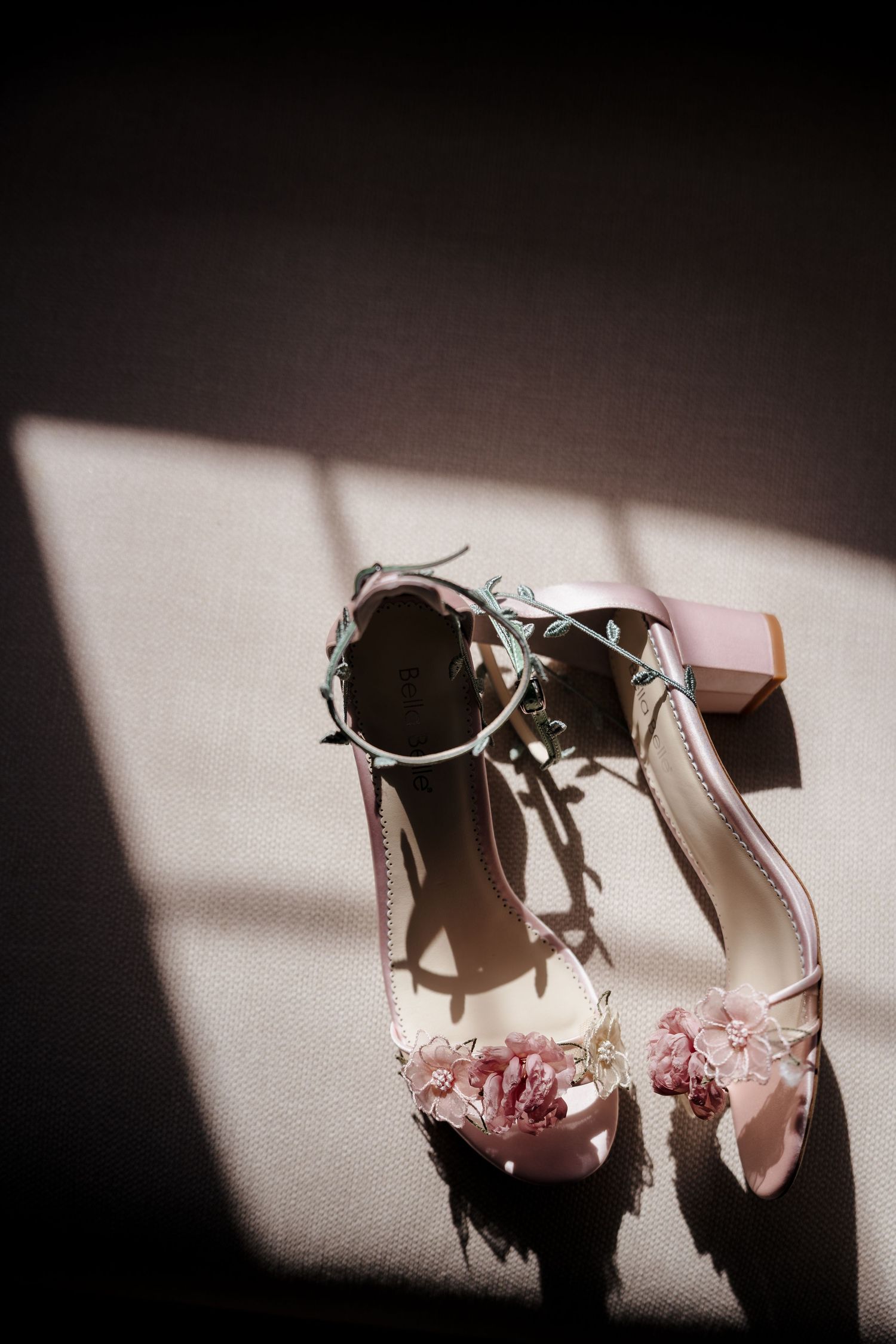 Elegant pink bridal heels with floral embellishments and ankle straps casting shadows in natural window light.