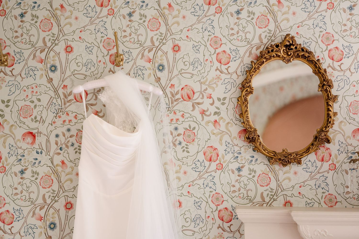 White wedding dress hanging on floral wallpaper beside ornate gold mirror in elegant bridal suite.