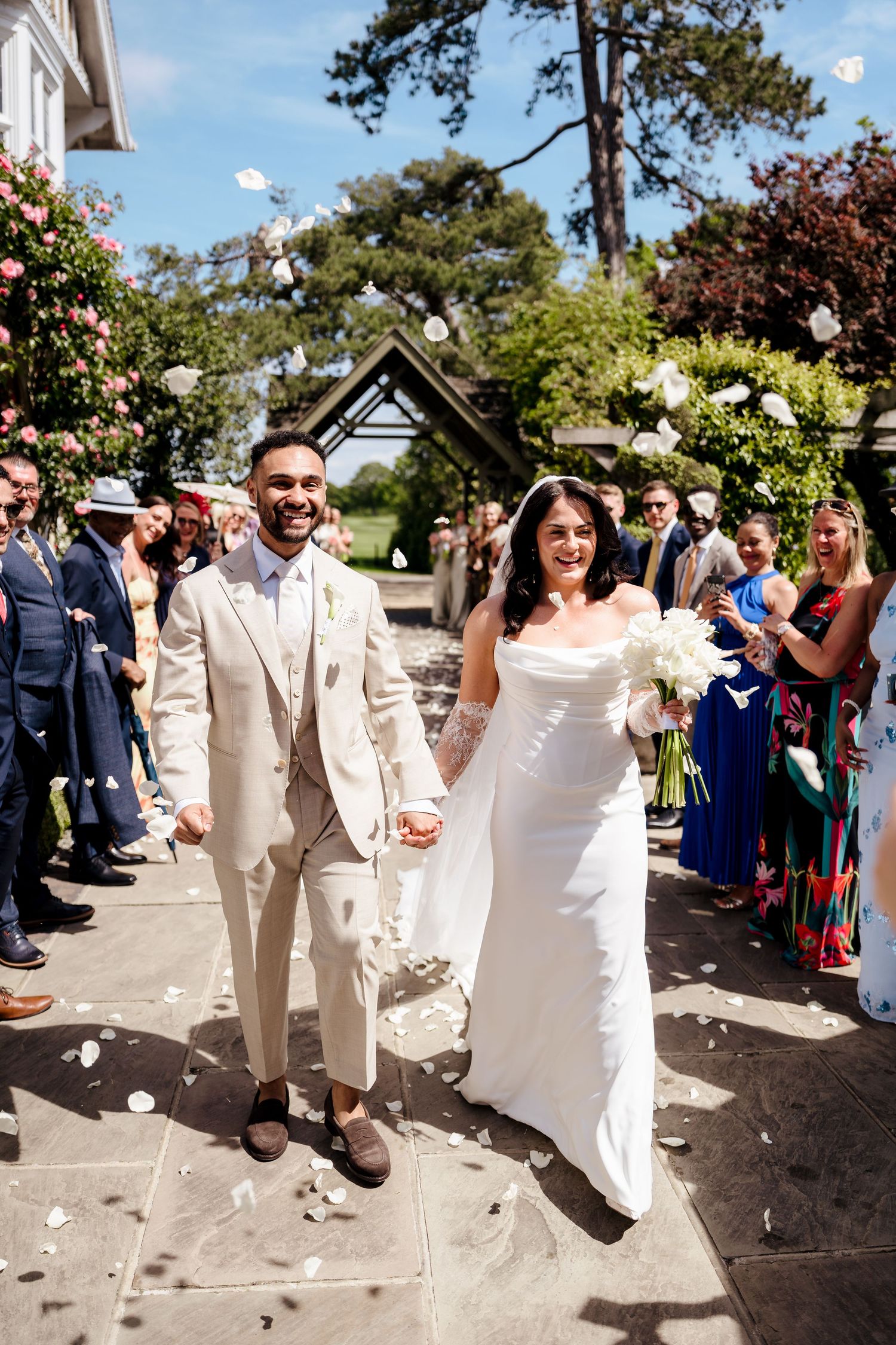 Bride and groom walk through confetti shower with smiling guests lining the path at outdoor wedding ceremony.