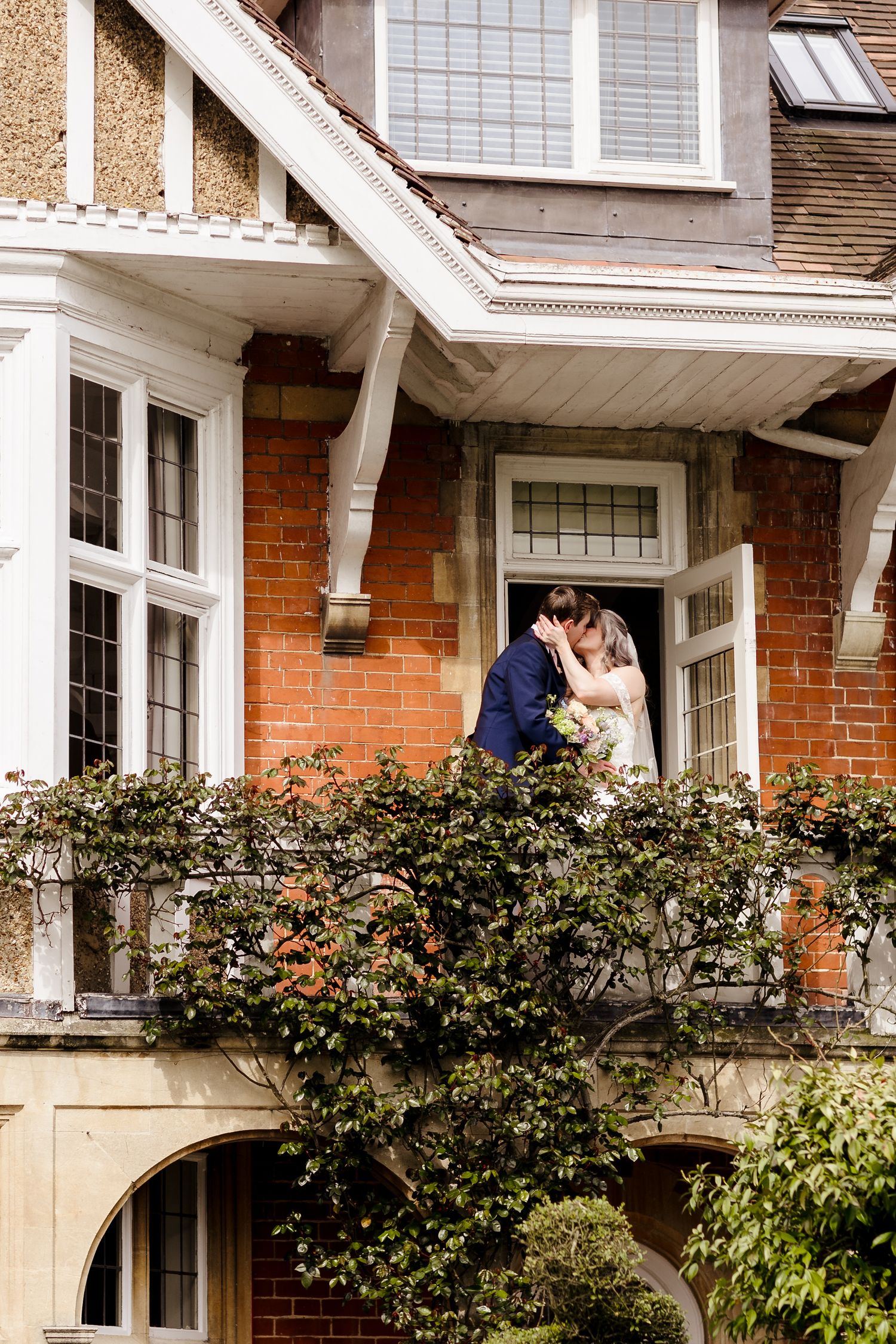 Bride and groom sharing a kiss on ivy-covered balcony of a classic red brick English country house venue.