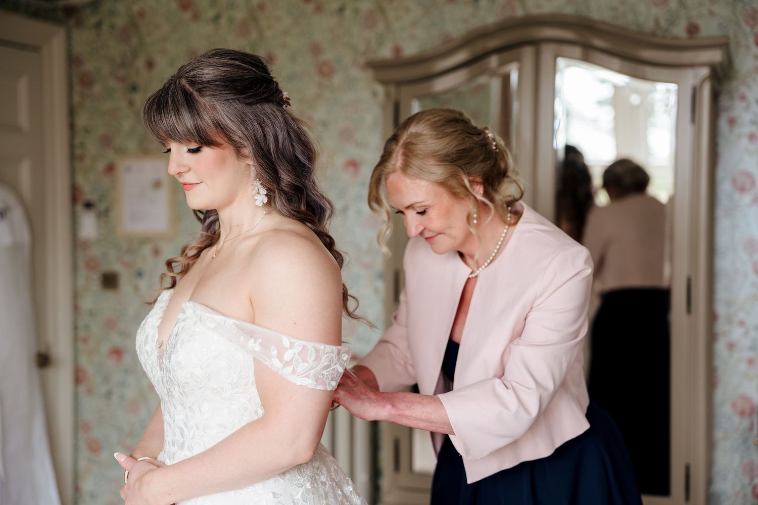 Mother helping bride into off-shoulder lace wedding dress in a floral wallpapered bridal suite.