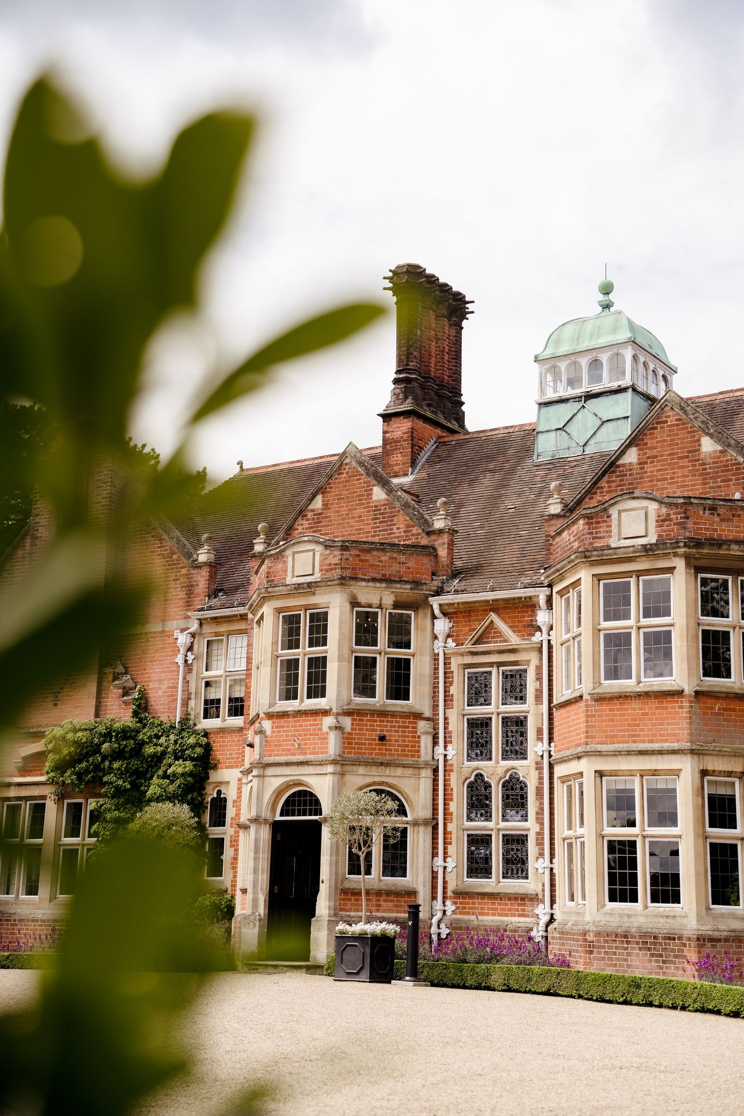 Grand red brick English manor house with ornate windows, copper dome, and manicured gardens under cloudy skies.