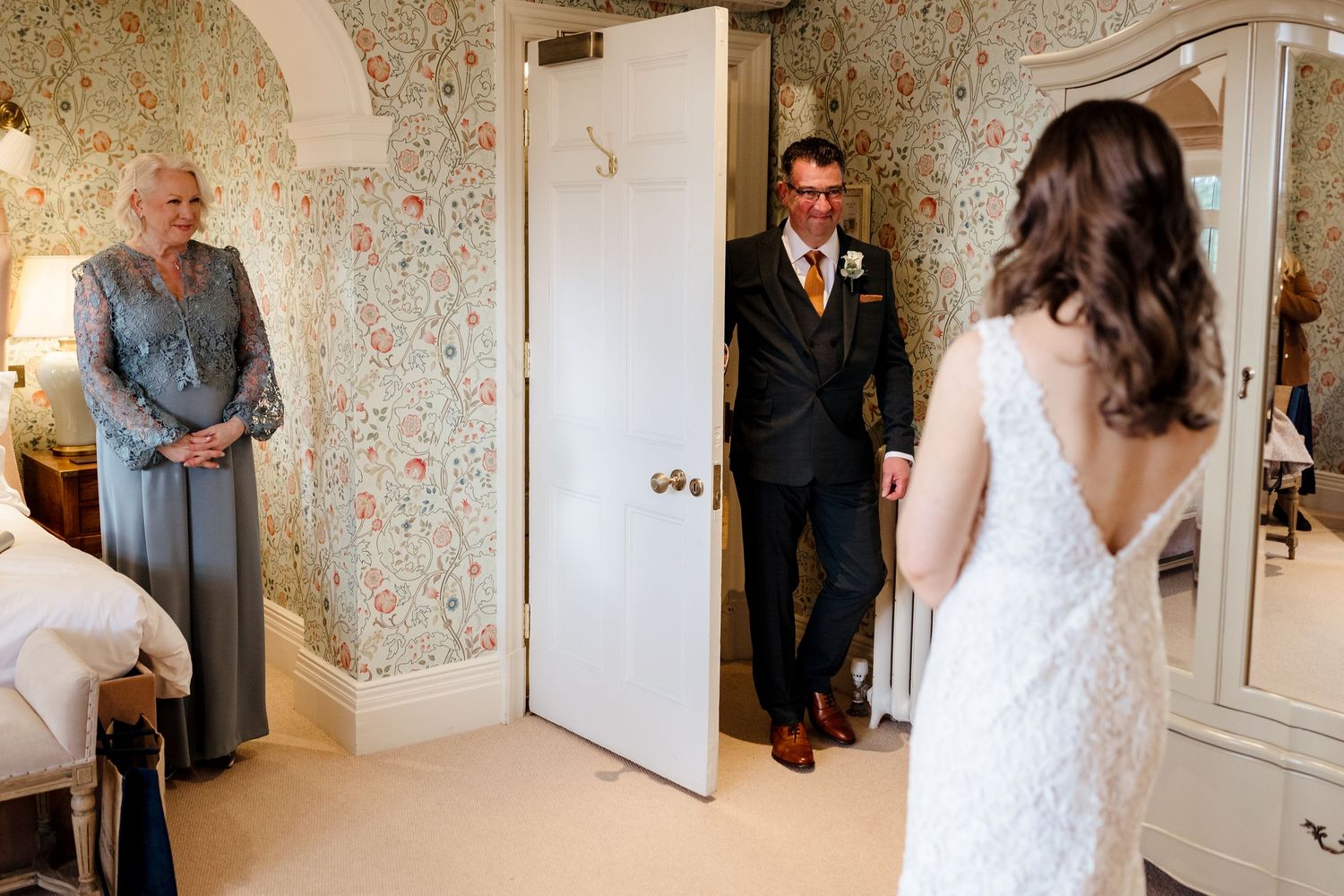 Groom in navy suit sees bride in white lace gown for first time, as older woman watches in floral wallpapered room.