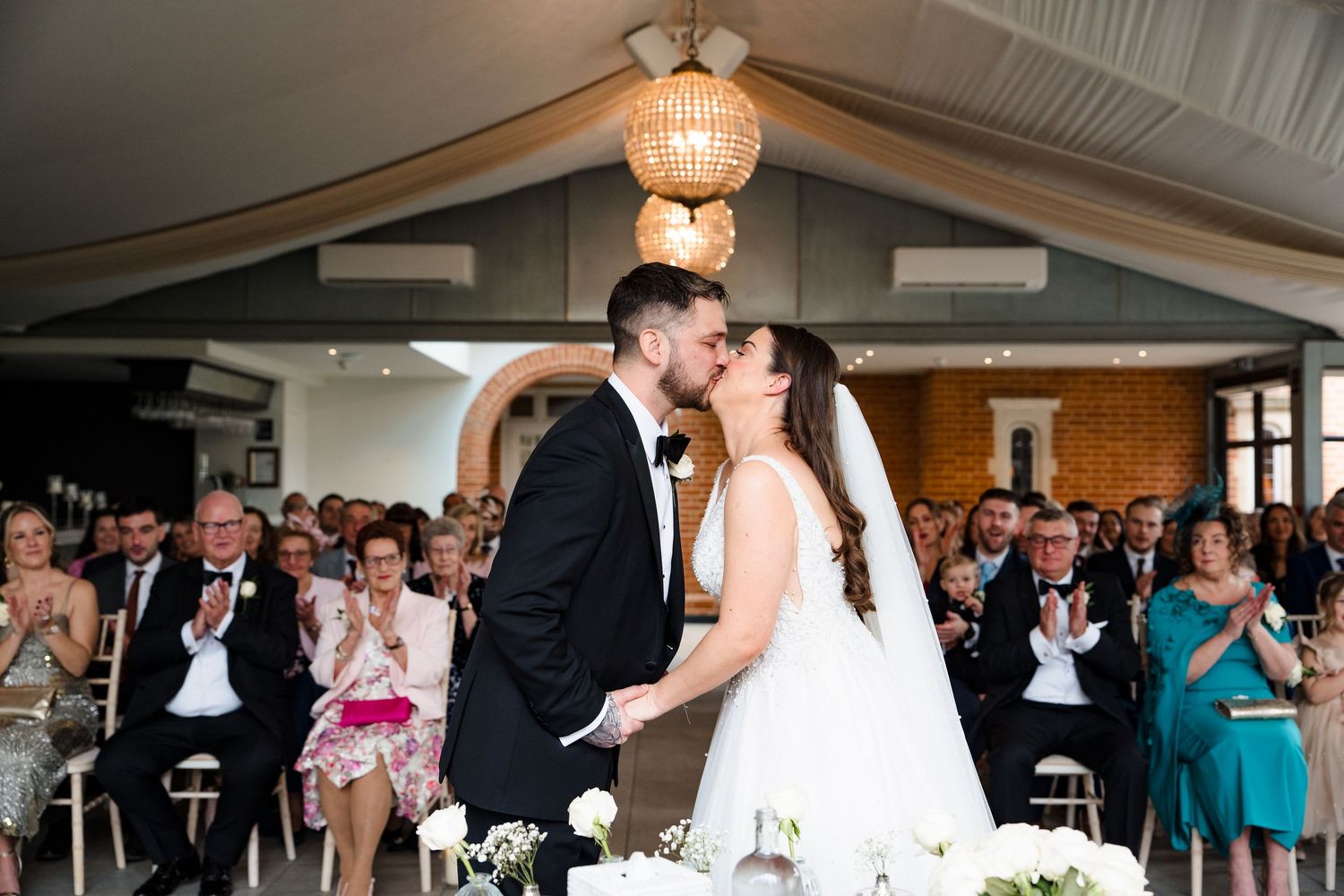 Newlyweds share their first kiss at the altar as wedding guests applaud in a bright elegant venue.