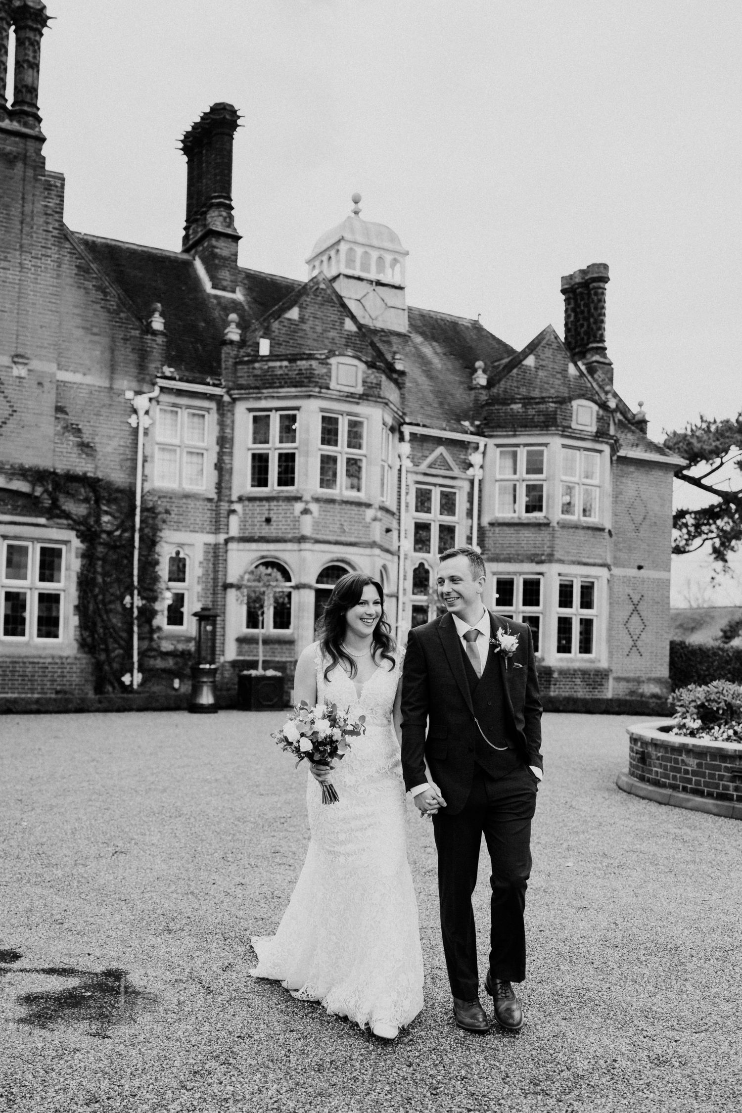 Black and white photo of bride and groom holding hands in front of a grand Victorian manor house wedding venue.